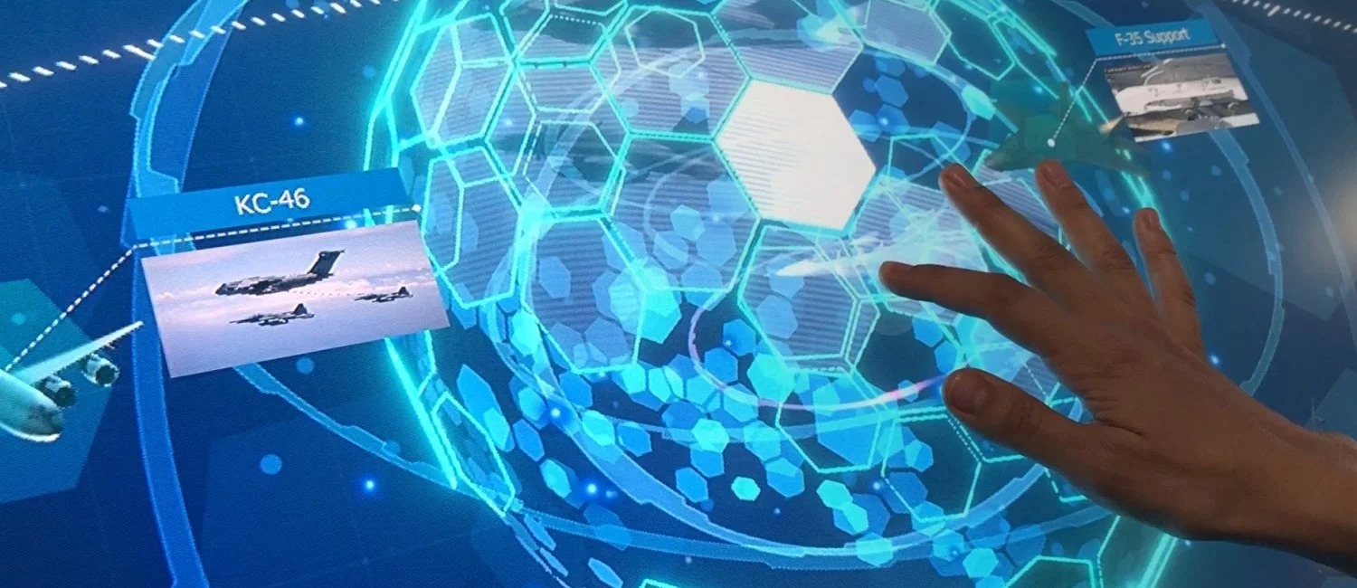A person's hand is touching a futuristic blue digital interactive display with holographic hexagon patterns, images of airplanes labeled KC-46 and F-35 support, and various technical graphics.