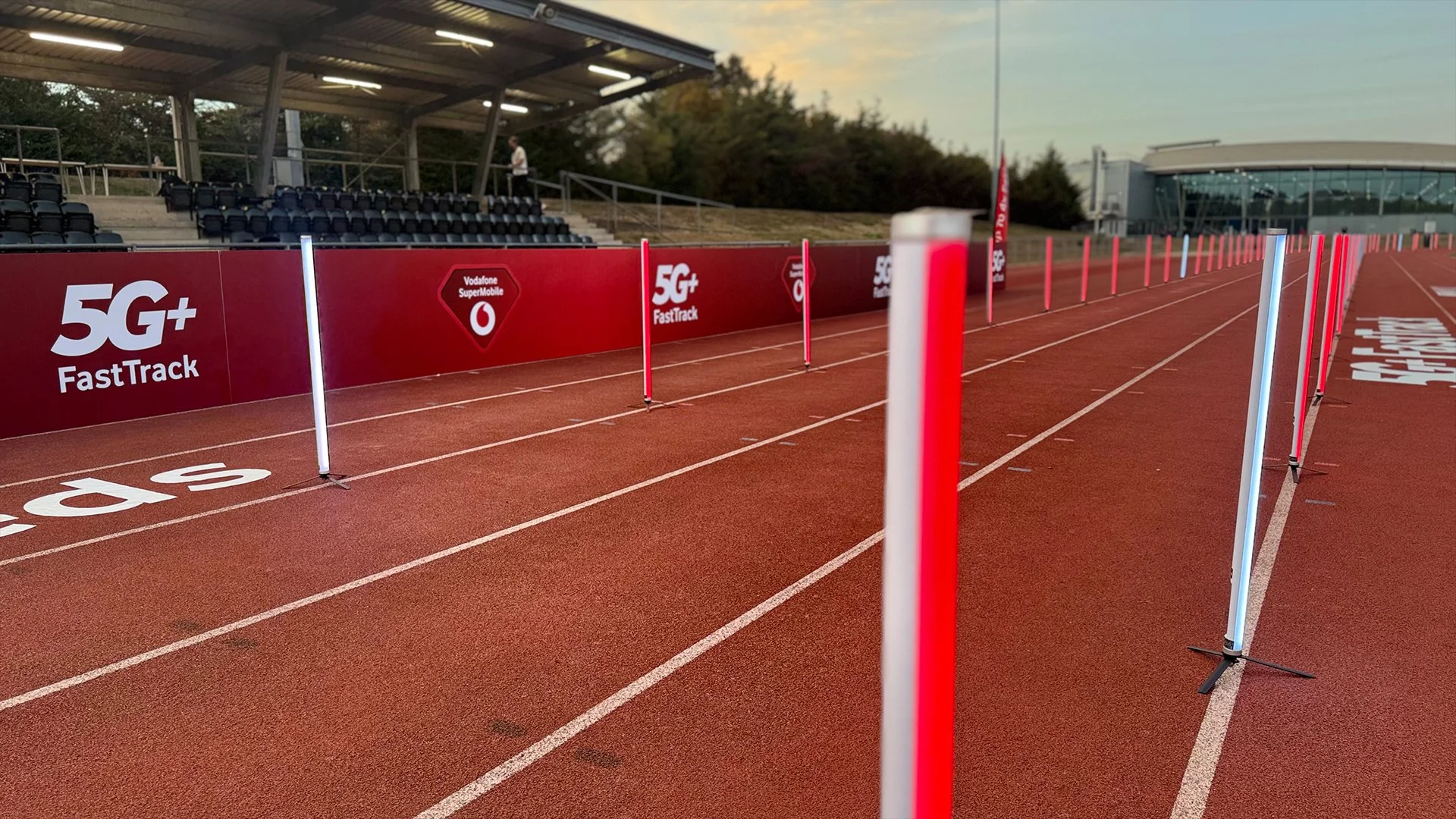 Empty outdoor running track with hurdles and promotional banners for 5G+ FastTrack and Vodafone SuperMobile, with a covered seating area and trees in the background.