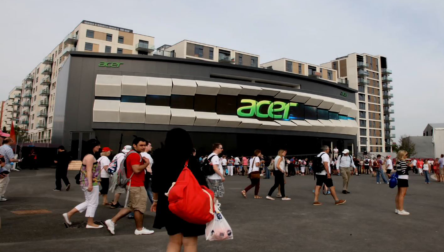 Crowd of people walking outside a modern building with an illuminated Acer logo.