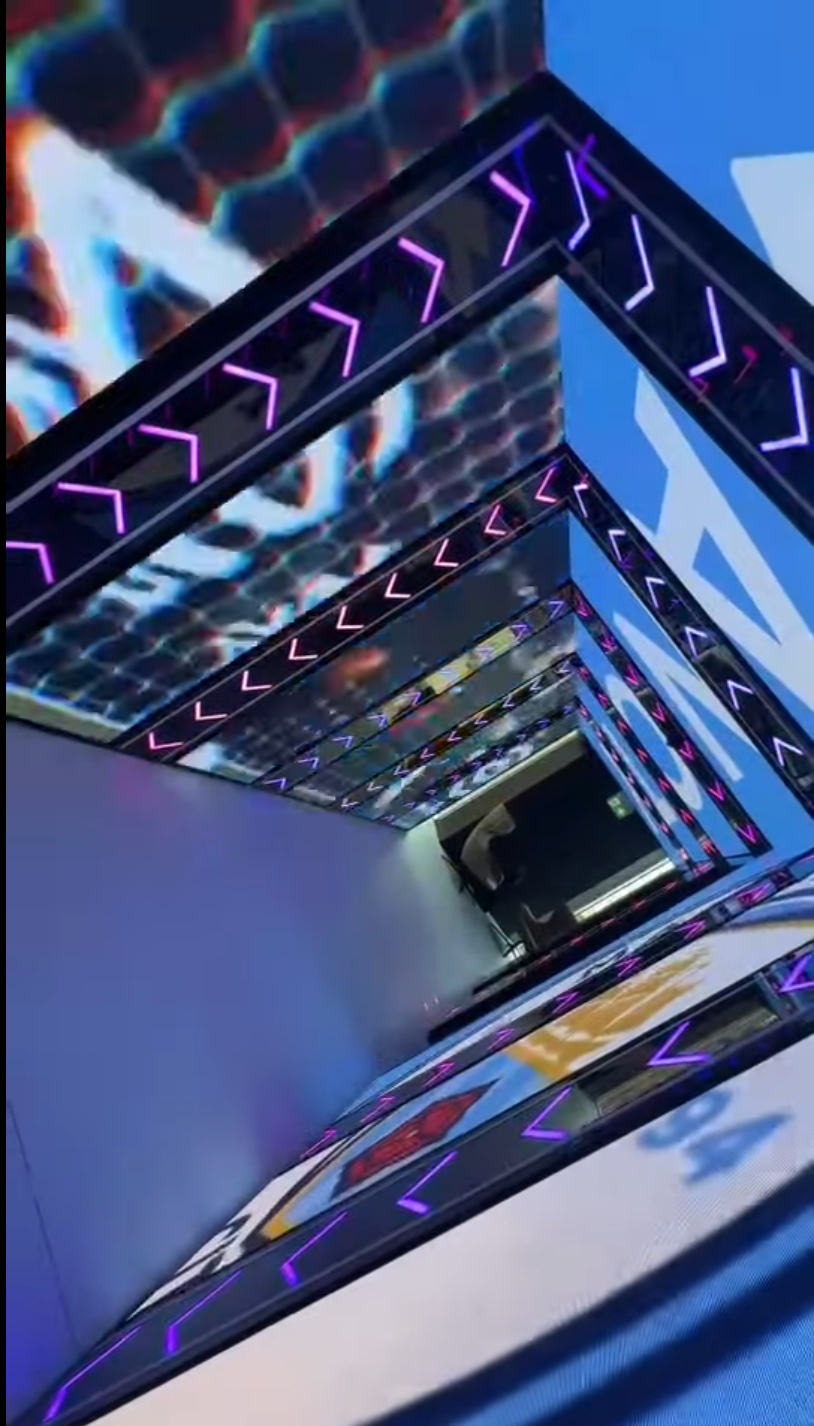 an indoor corridor with futuristic design featuring neon lights in purple and pink colors, geometric ceiling patterns, and a reflective surface on the floor.