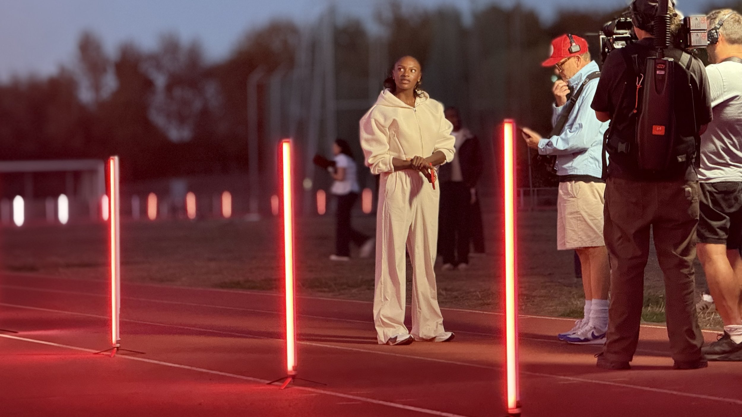 A woman in beige tracksuit standing on a red track surrounded by vertical red glowing poles, with a group of people, some wearing headsets, in the background during dusk.