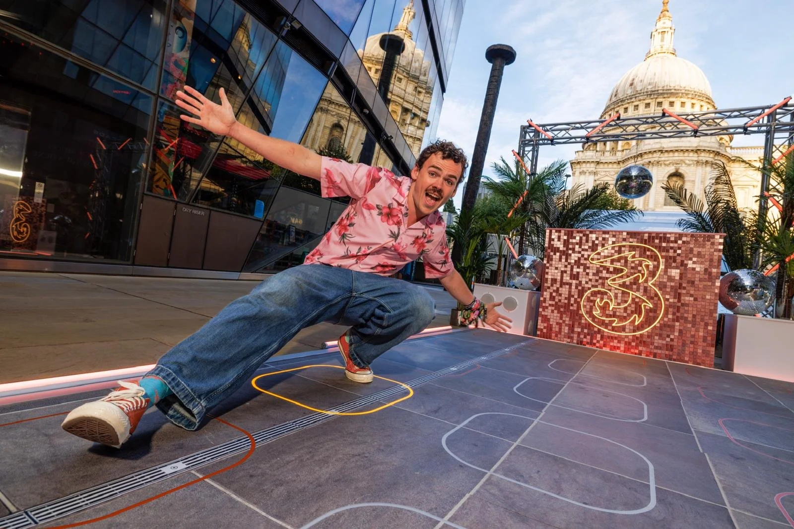 A man in a pink floral shirt and jeans crouches on a painted court on a city street, smiling and making a lively gesture. Behind him is a colorful neon sign with the number '3', palm plants, and a historic building with a dome, suggesting a festive outdoor event.