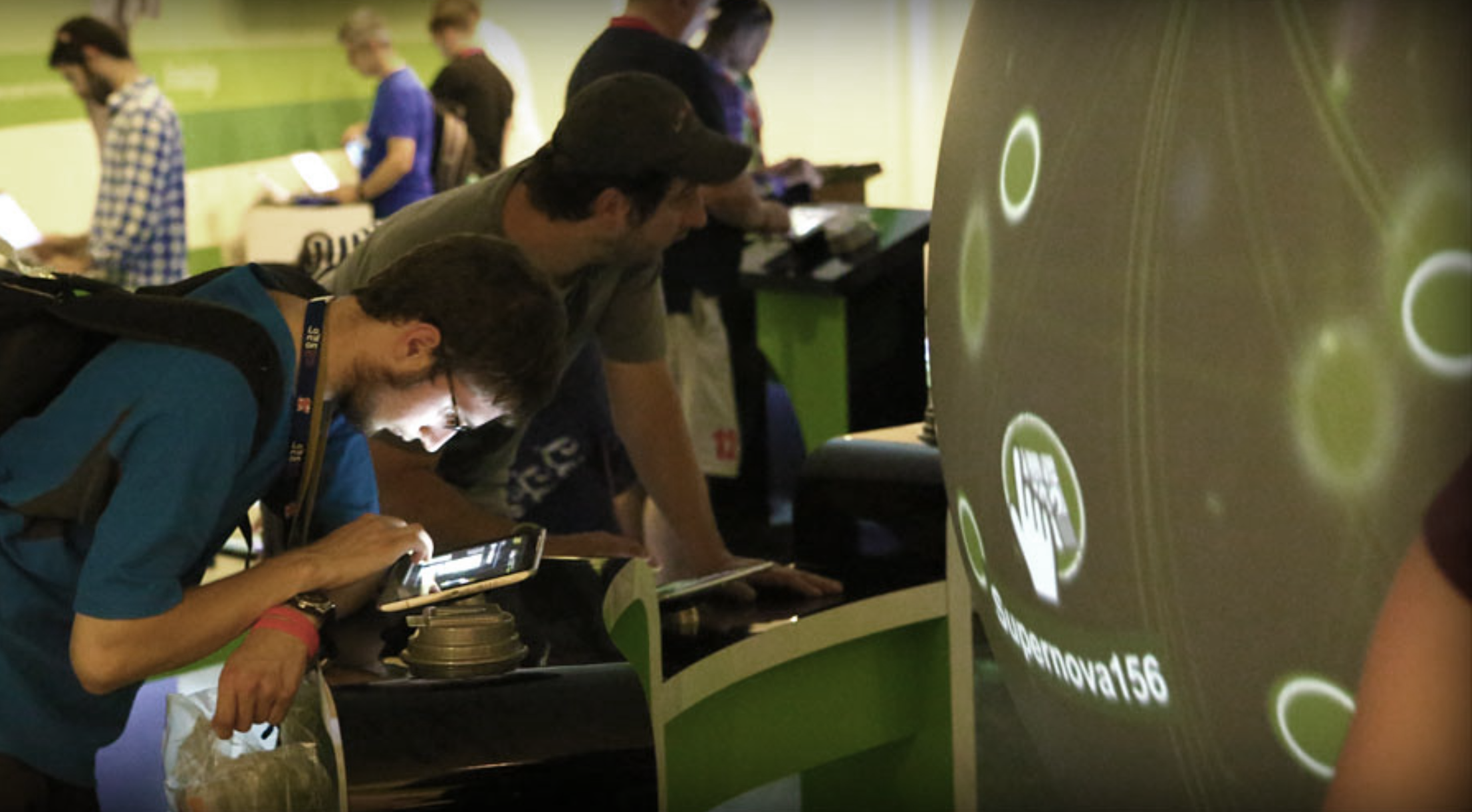 People at a technology or gaming expo, with two individuals intensely focused on a digital device, and others interacting with screens in the background.