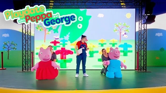 A stage with a large screen displaying a colorful cartoon background. On the stage, there are two people dressed as Peppa Pig characters, Playdate with Peppa and George Theatre Legoland New York Parachute studios