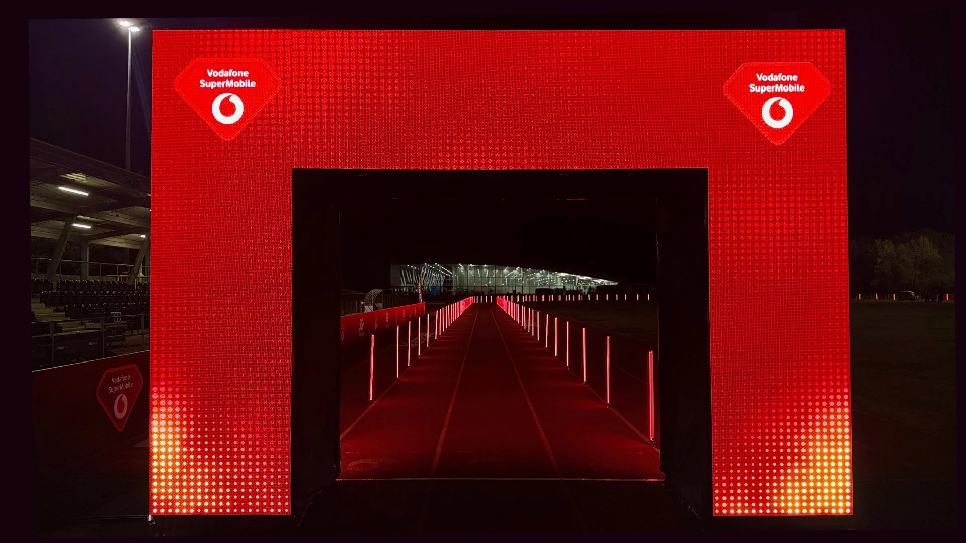 Nighttime scene of a large red digital structure with Vodafone SuperMobile logos at top corners, creating a tunnel-like entrance with illuminated red lights along pathways.
