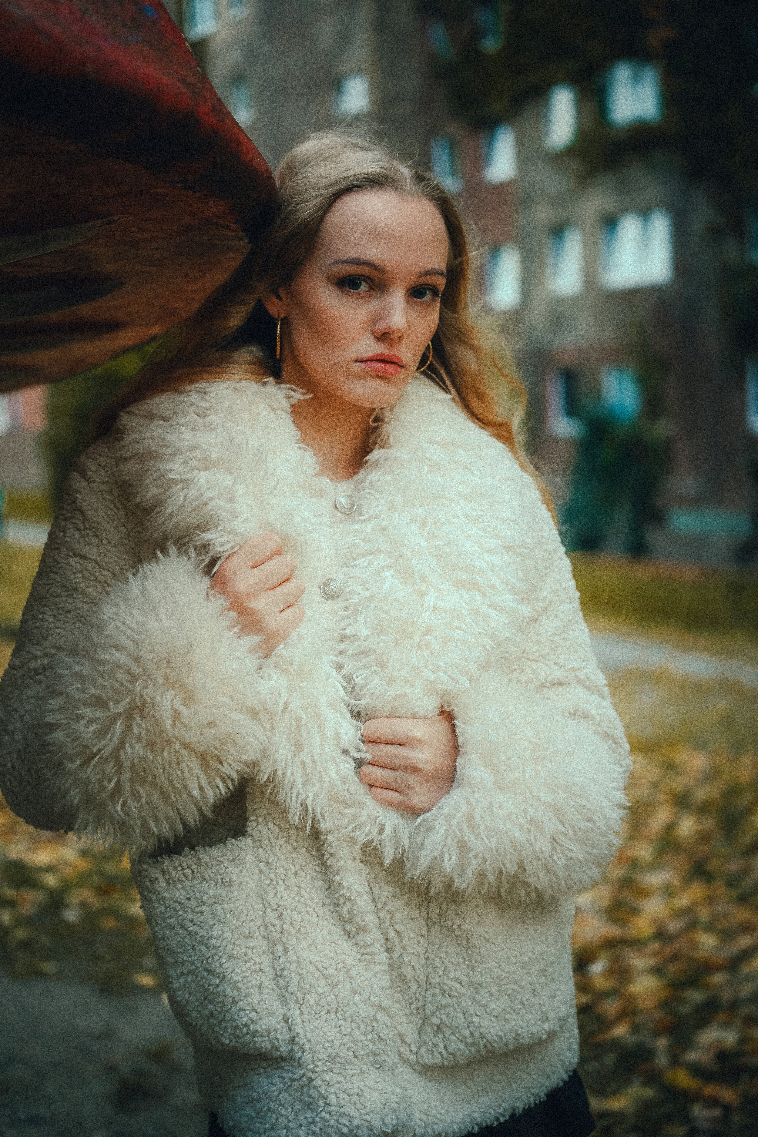 Eine junge Frau mit langen, lockigen Haaren trägt eine weiße, fellartige Jacke und steht im Freien vor einem Hintergrund mit Bäumen und Gebäuden. Sie sieht ernst aus und hält die Jacke mit beiden Händen fest.