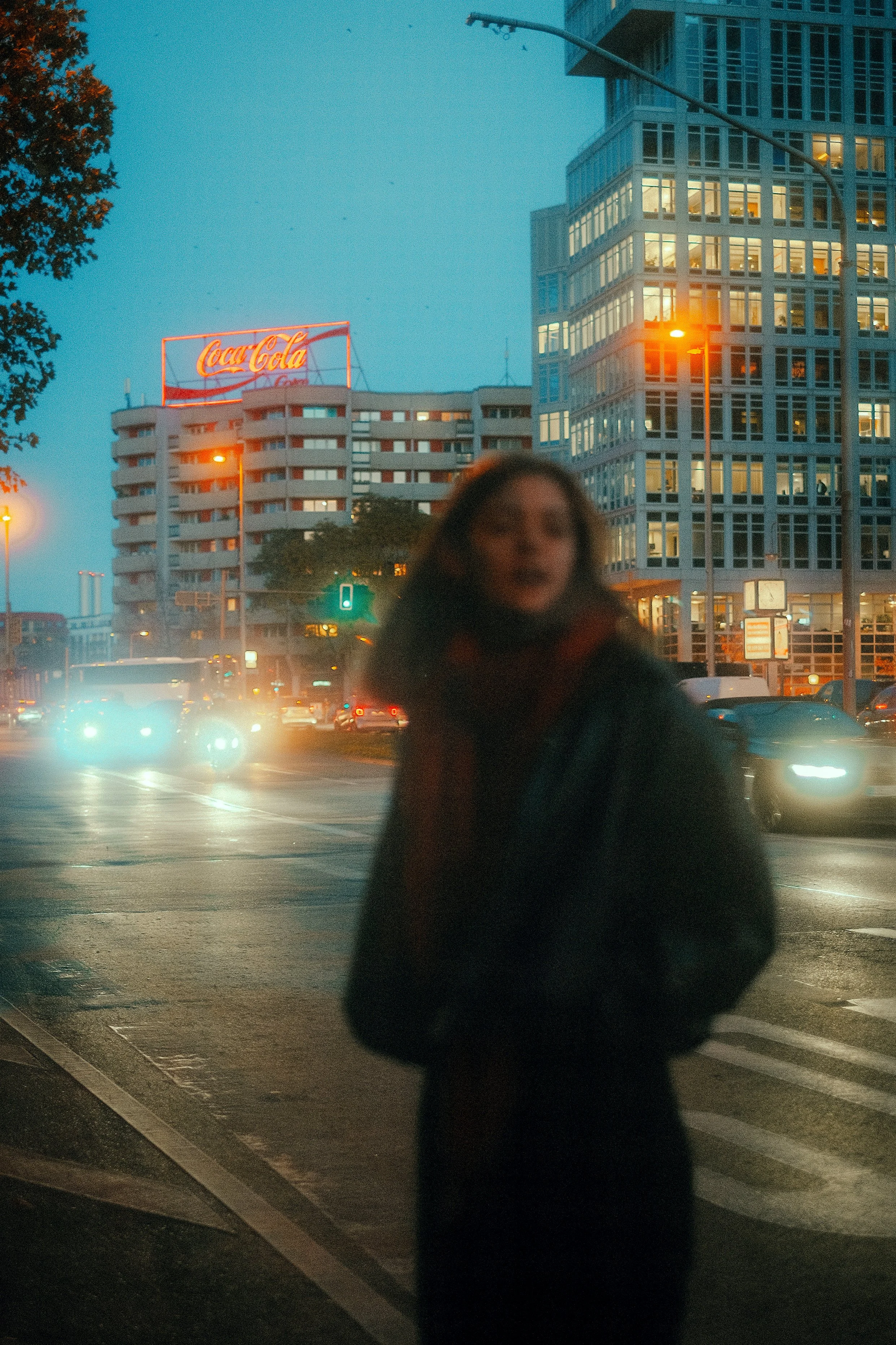 Eine unscharfe Frau steht an einer Kreuzung in einer urbanen Gegend bei Dämmerung, im Hintergrund sind mehrere Gebäude, ein Coca-Cola-Schild und Autos mit eingeschalteten Scheinwerfern sichtbar.