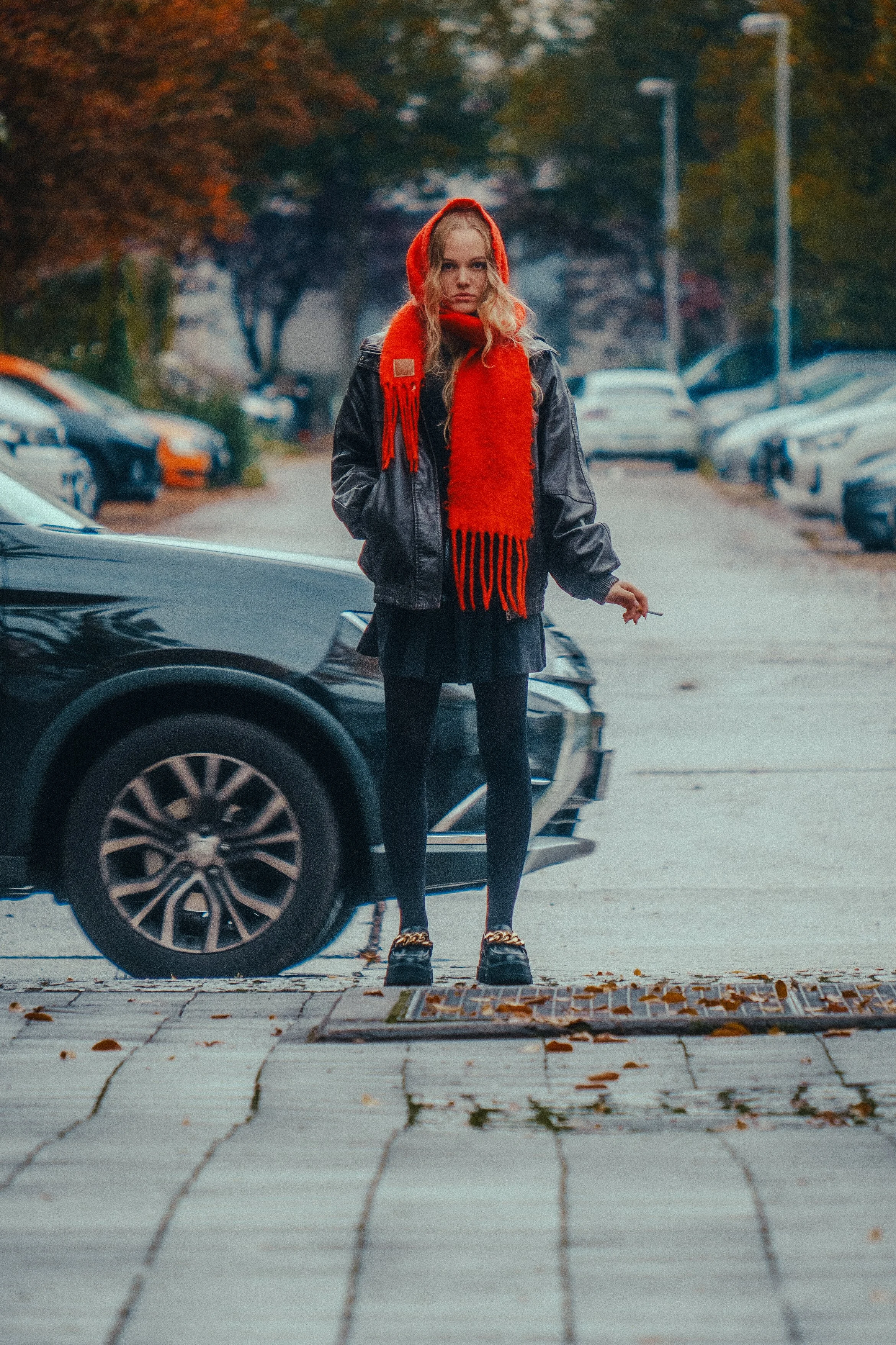 Junge Frau mit roten Schal und Kapuze steht auf Gehweg im Parkhaus, umgeben von geparkten Autos, im Herbst, düstere Stimmung