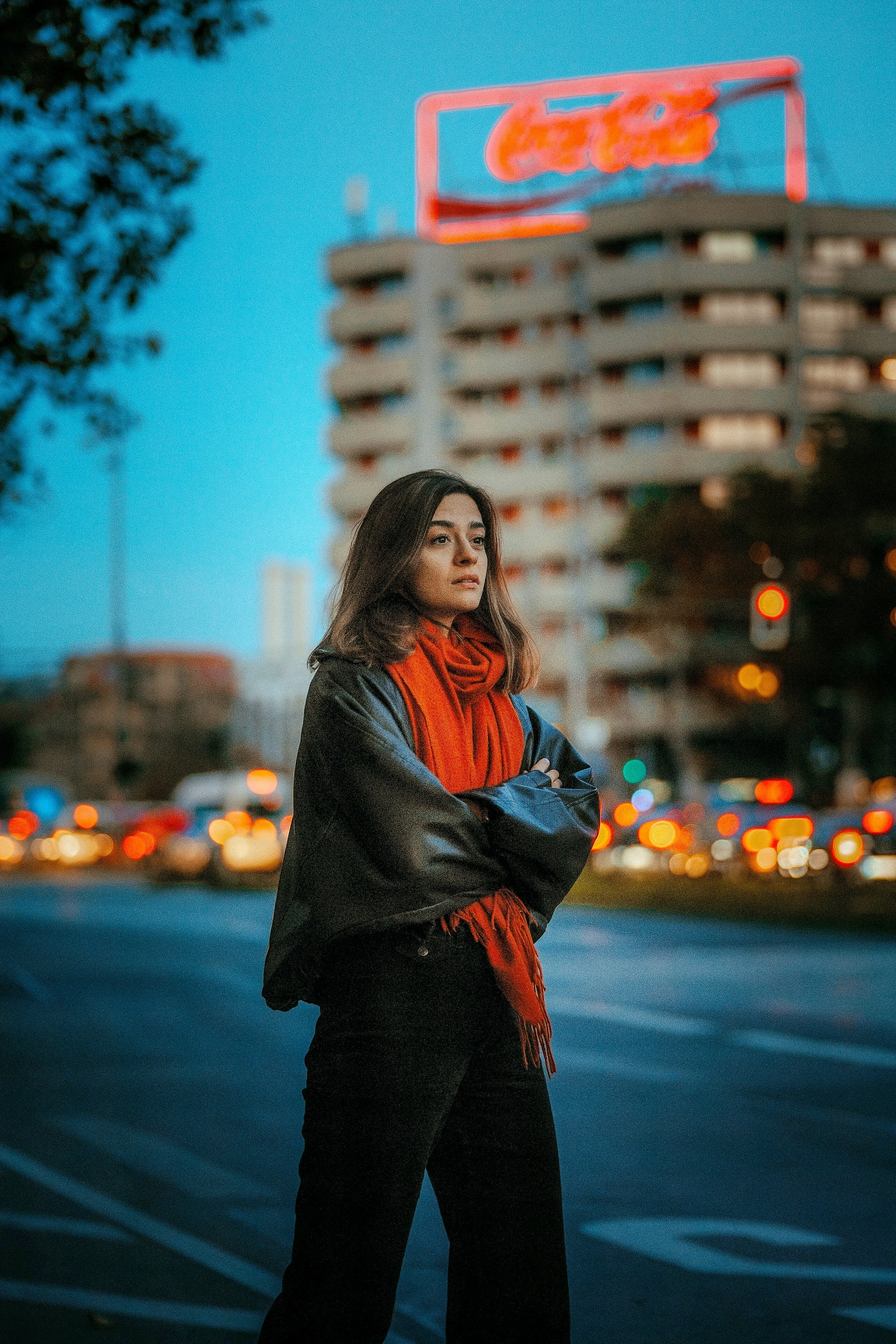 Eine Frau steht in einer Stadt bei Dämmerung, umgeben von Verkehrslicht und einem Hochhaus mit einem leuchtenden Neon-Schild im Hintergrund.