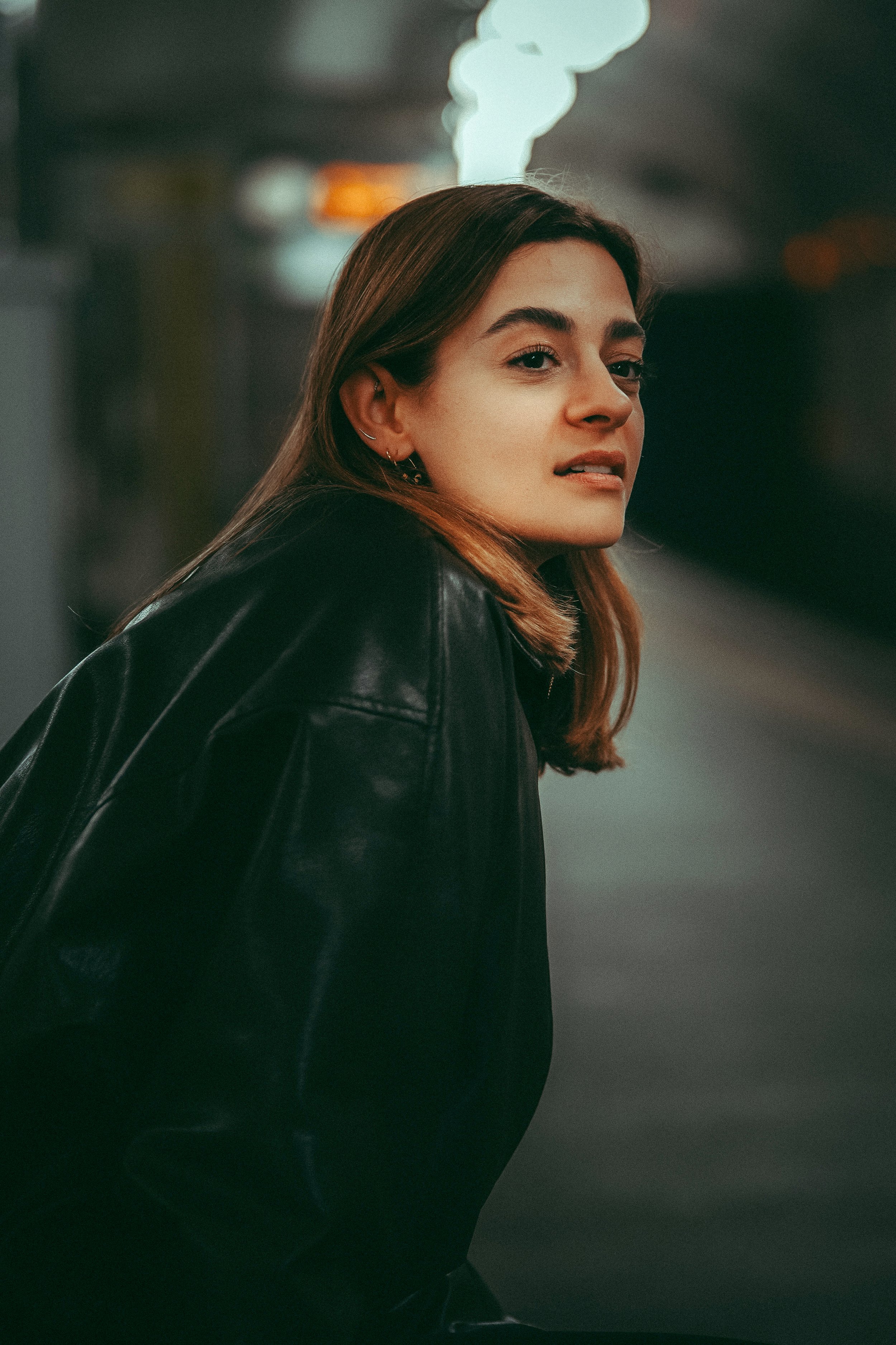 Junge Frau mit braunen Haaren in schwarzer Lederjacke, blickt versonnen, im Hintergrund verschwommene Lichter, vermutlich in einer Stadt bei Nacht.