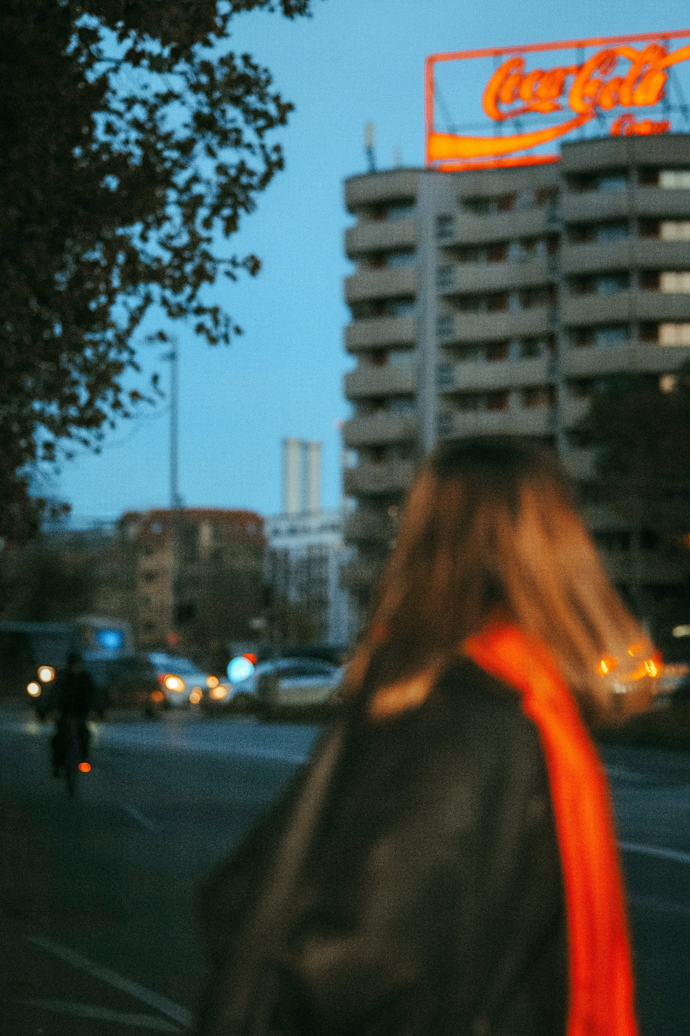 Eine Person mit braunen Haaren steht neben einer Straße bei Dämmerung, im Hintergrund sind Autos, ein Gebäude mit einem Coca-Cola Neon-Schild und ein Baum sichtbar.