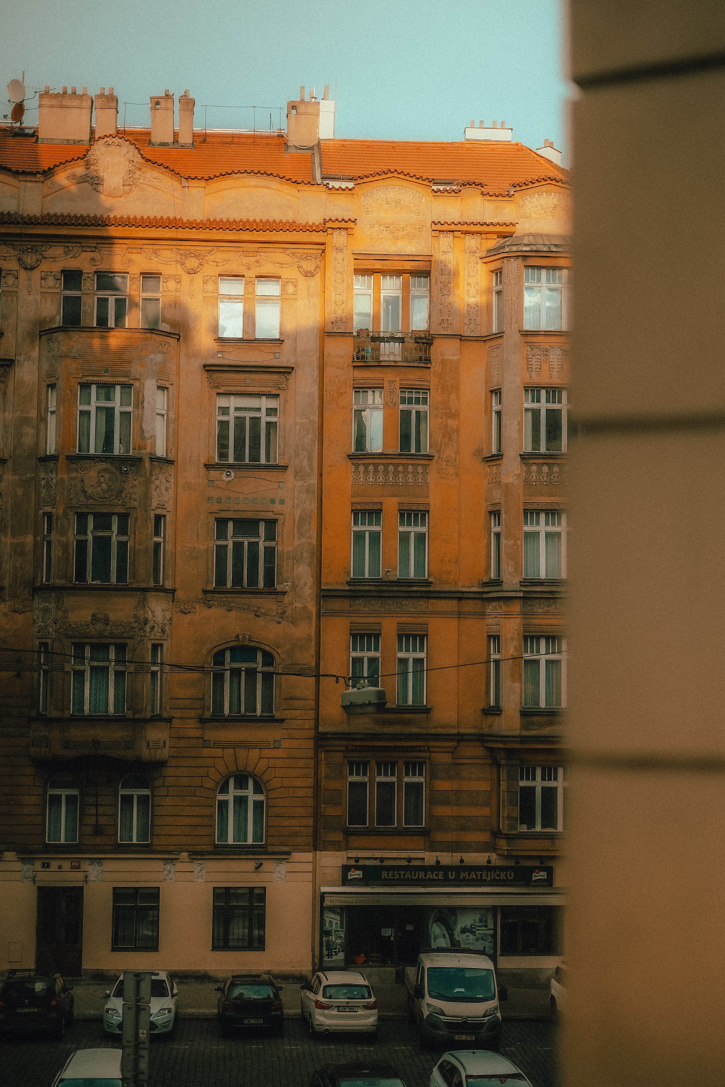 Ein altes Mehrfamilienhaus mit orangefarbenen Fassade und mehreren Fenstern, im unteren Bereich eine Kneipe namens 'Restaurace u Matějíčku', davor geparkte Autos.