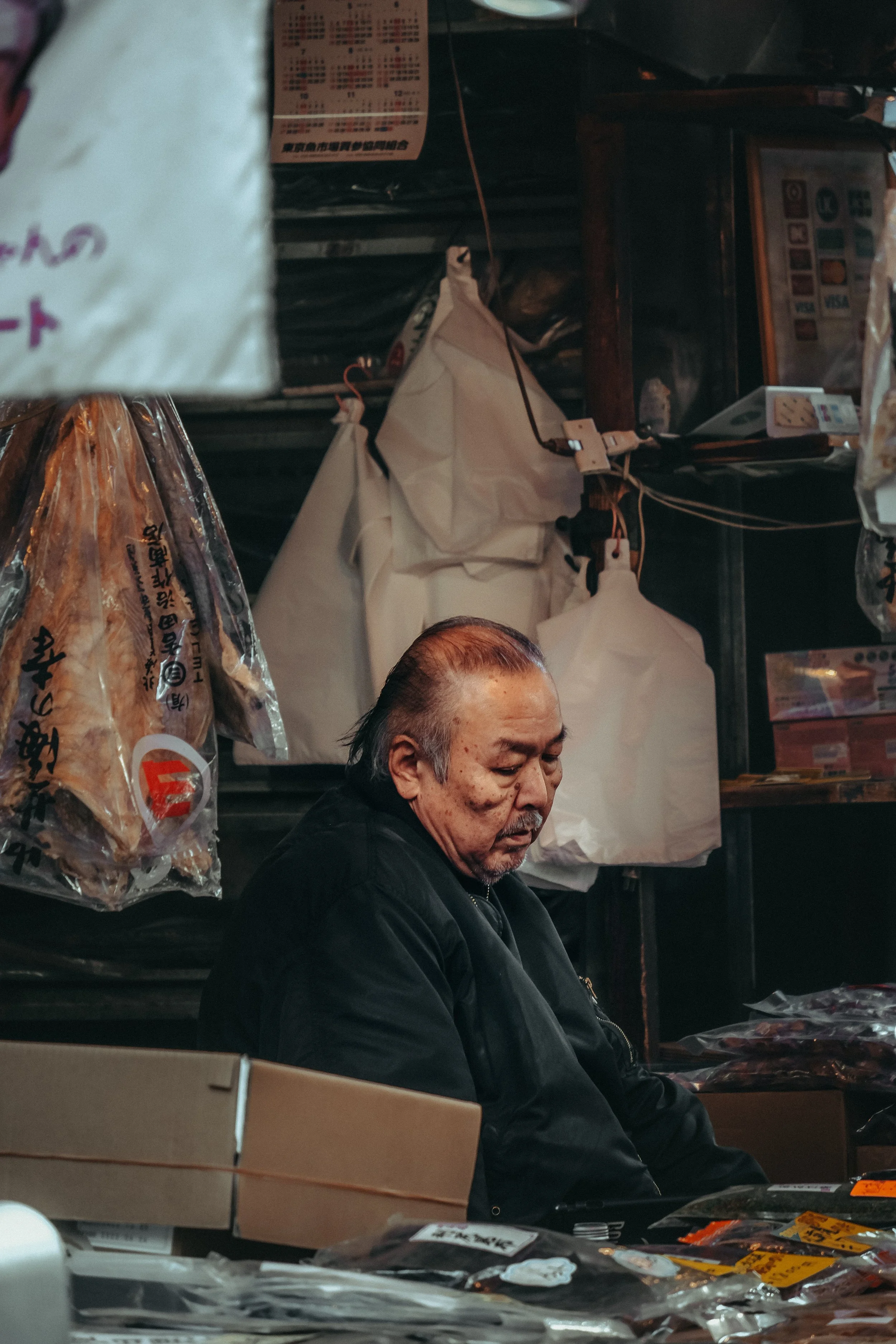 Älterer Mann sitzt in einem Marktstand, umgeben von verpackten Lebensmitteln und Verpackungen, mit weißen Tüten im Hintergrund und einem Verkaufsdisplay auf der rechten Seite.