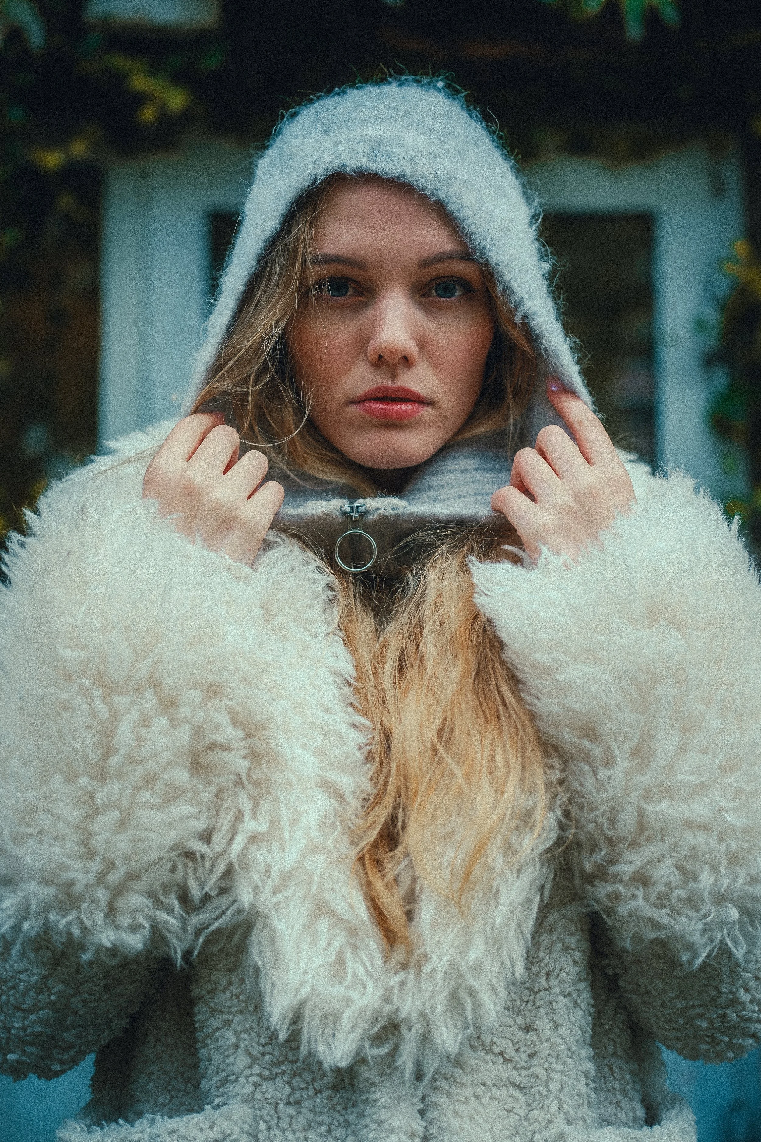 Nahaufnahme einer jungen Frau in warmer Kleidung, trägt einen weißen pelzigen Mantel und eine graue Kapuzenjacke, im Hintergrund ein Haus mit weißer Tür.