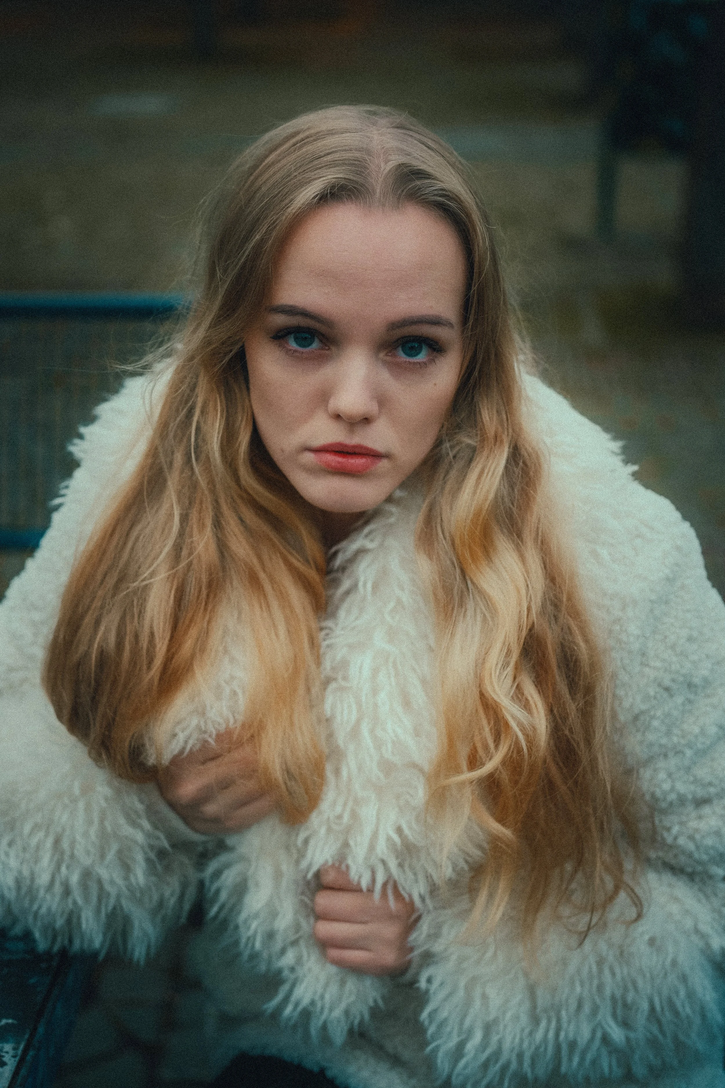 Nahaufnahme einer jungen Frau mit langen, lockigen blonden Haaren, blauen Augen und rotem Lippenstift, die eine weiße, plüschige Jacke trägt.