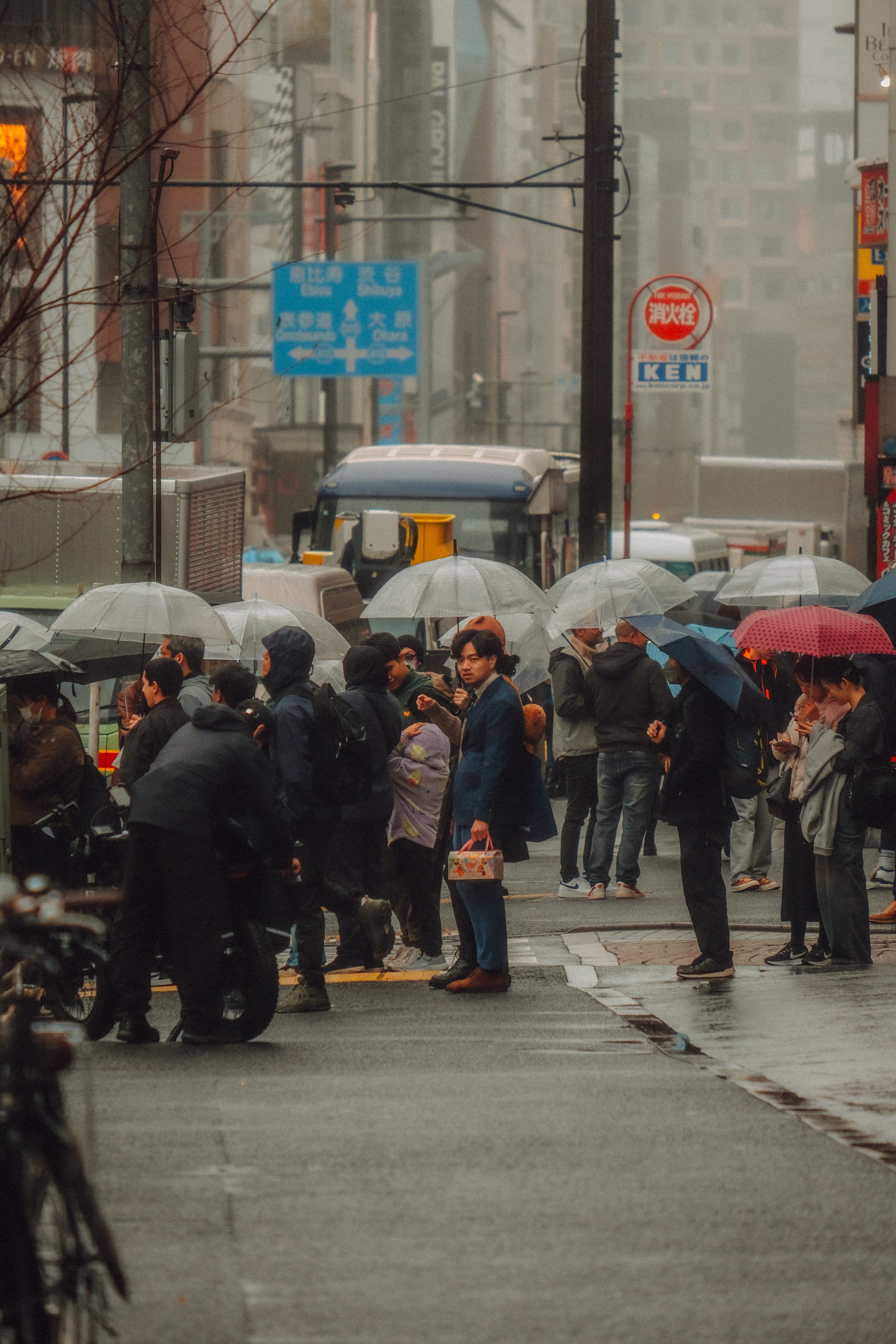 Menschen stehen bei Regen mit Regenschirmen an einer Straßenecke in einer urbanen Umgebung.