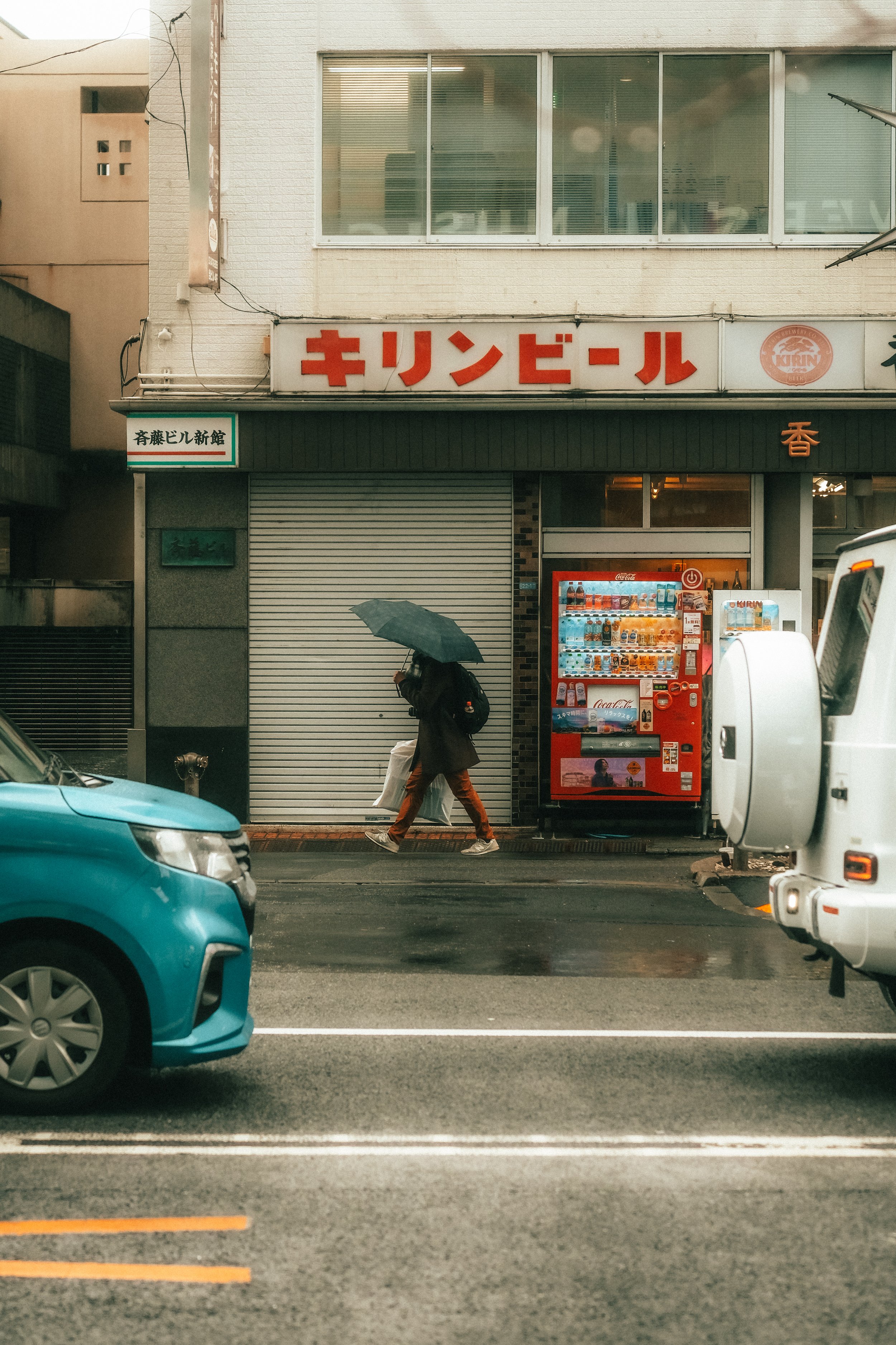 Ein Mann mit Regenschirm läuft an einer Geschäftefassade vorbei, auf der ein Getränkestand mit Getränkemaschine und mehreren Getränken steht, während es regnet.