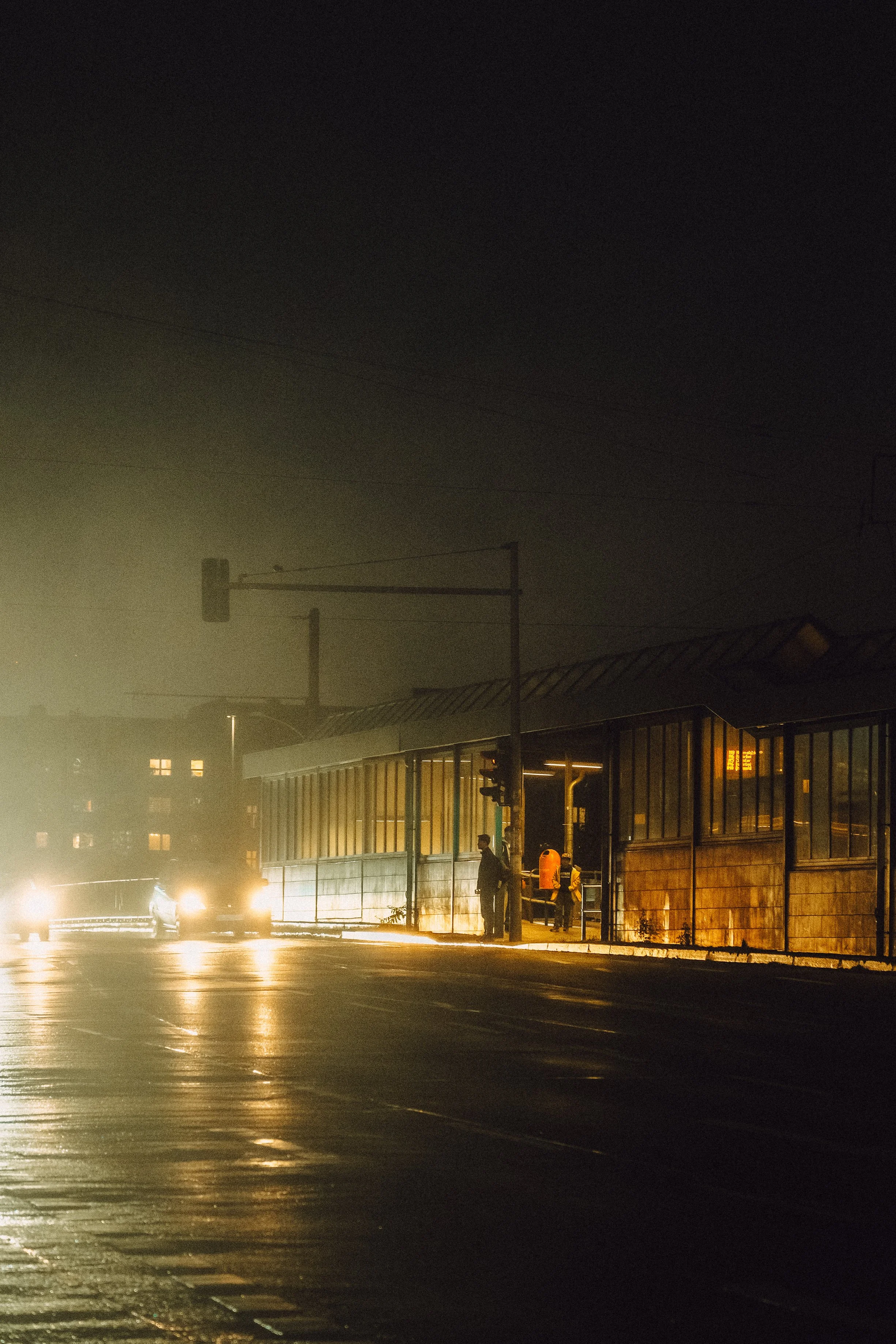 Nachtaufnahme an einer Station mit zwei Polizisten im Einsatz, bei schlechter Sicht durch Nebel oder Rauch, beleuchtet von Straßenlaternen und Fahrzeuglichtern.