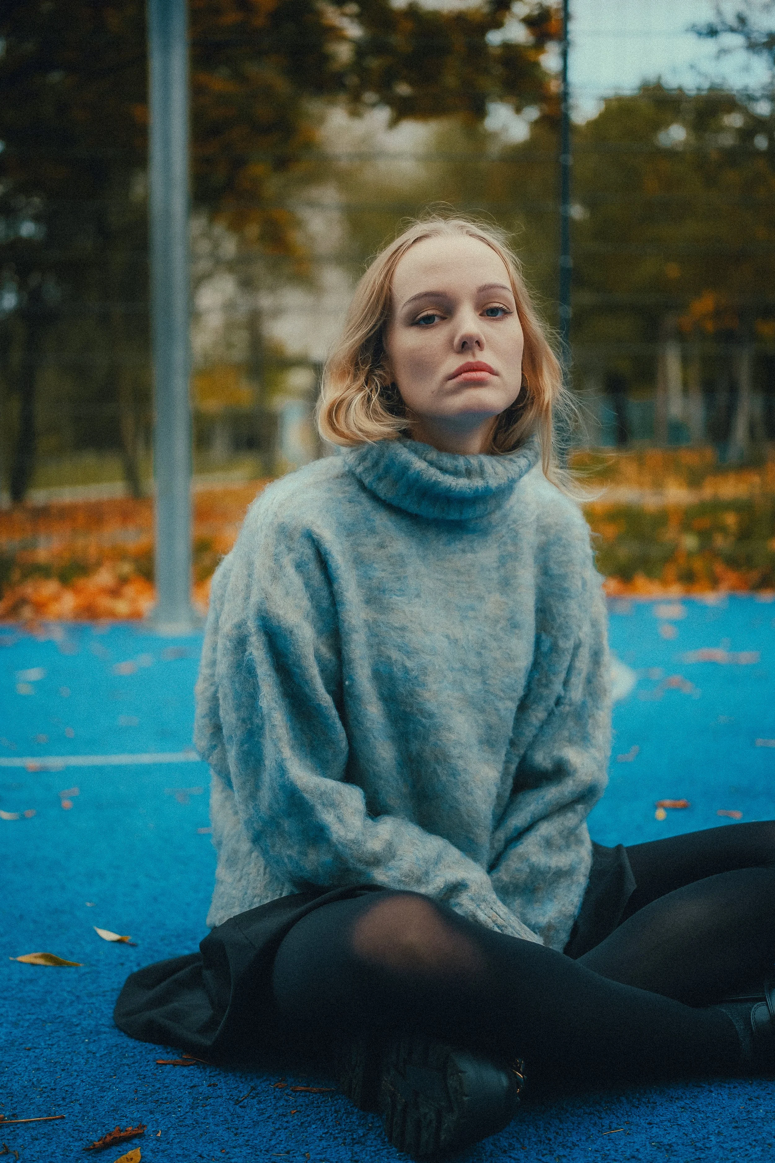 Ein Mädchen sitzt auf einem blauen Tennisplatz im Freien, trägt einen grauen Rollkragenpullover und schwarze Strumpfhosen, umgeben von herbstlichen Herbstblättern.