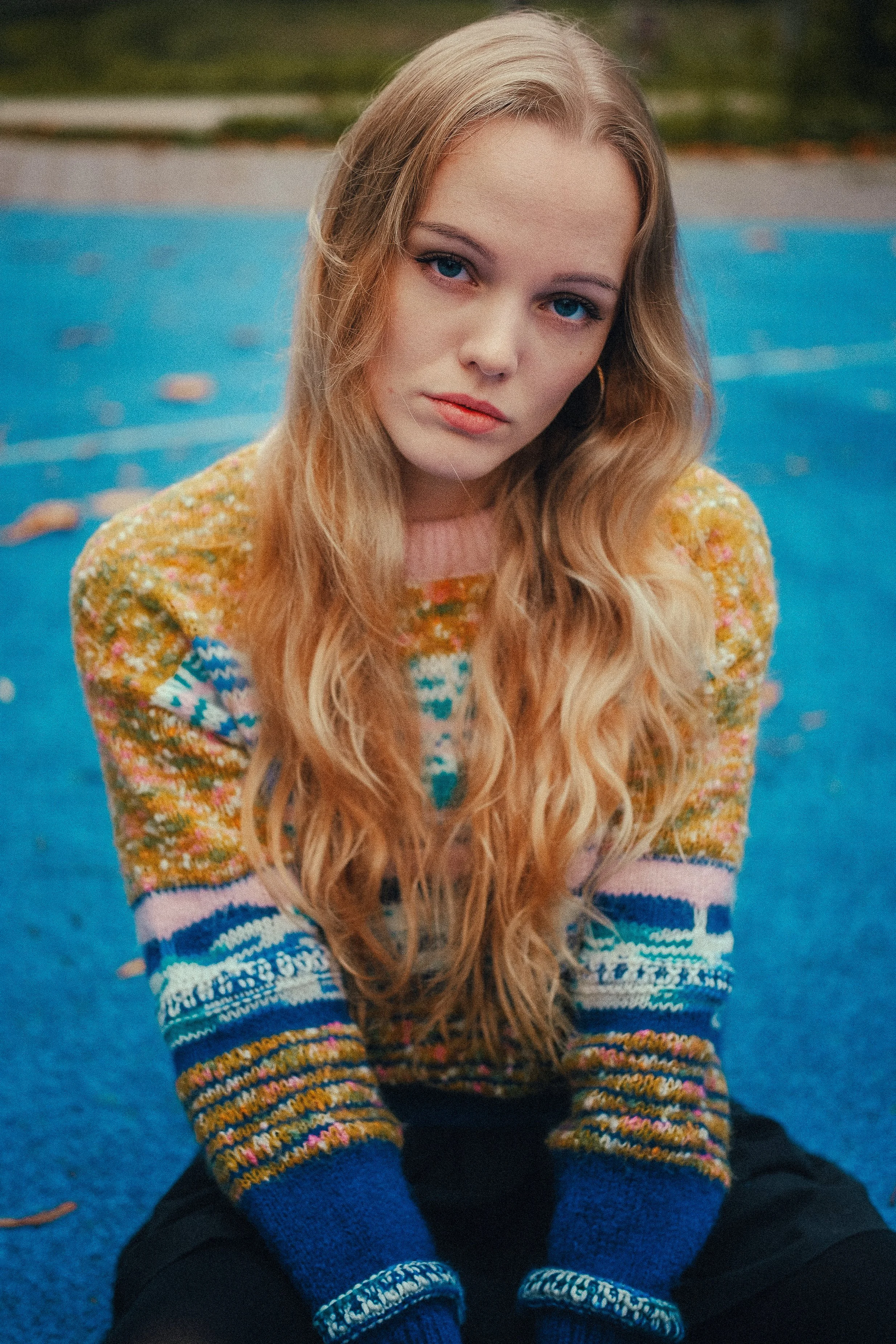Junge Frau mit langen, lockigen blonden Haaren trägt einen bunten, gemusterten Pullover und sitzt auf blauer Oberfläche, vermutlich im Freien.