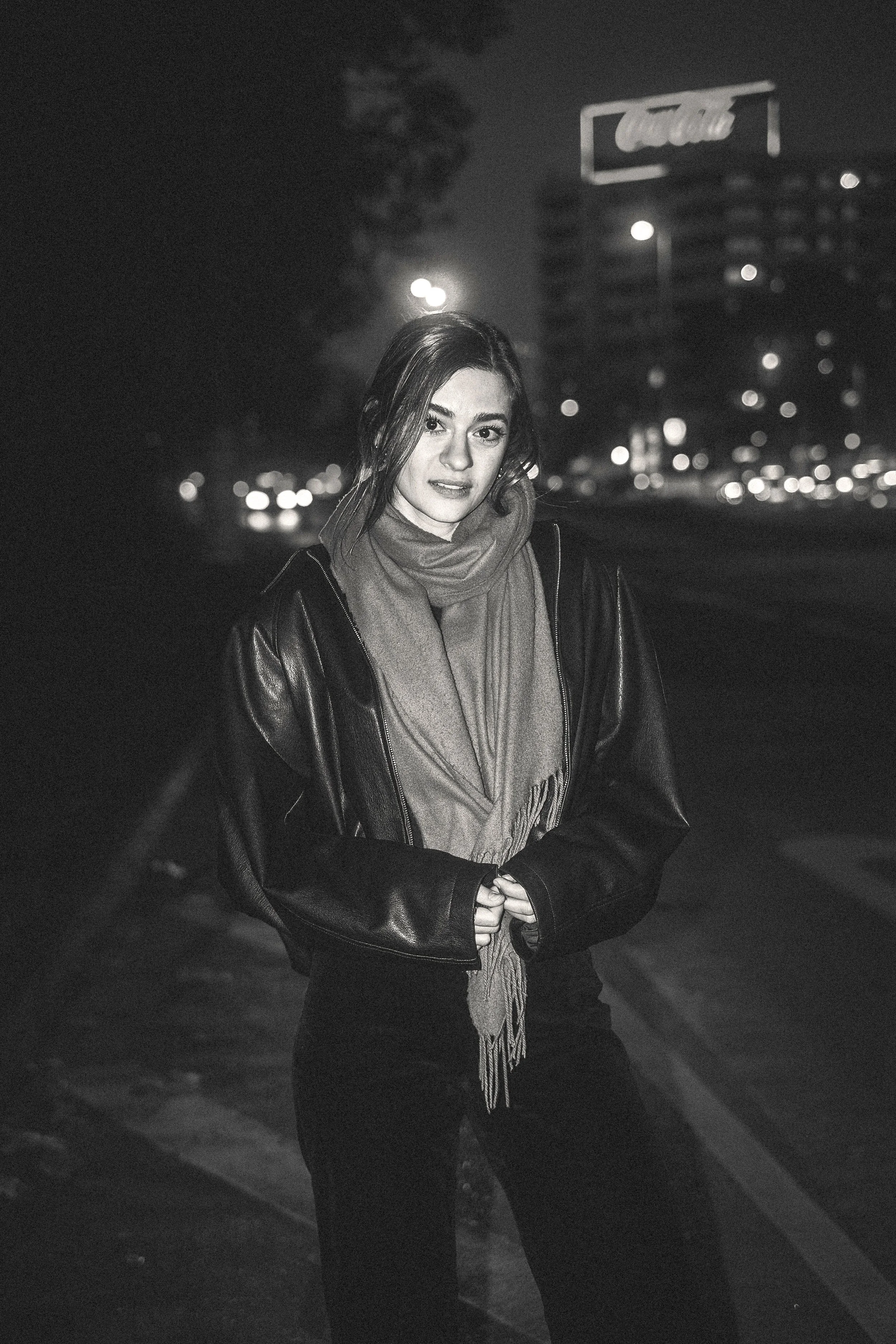 Frau in Jacke und Schal steht nachts an einer Straßenkreuzung mit Lichtern im Hintergrund.