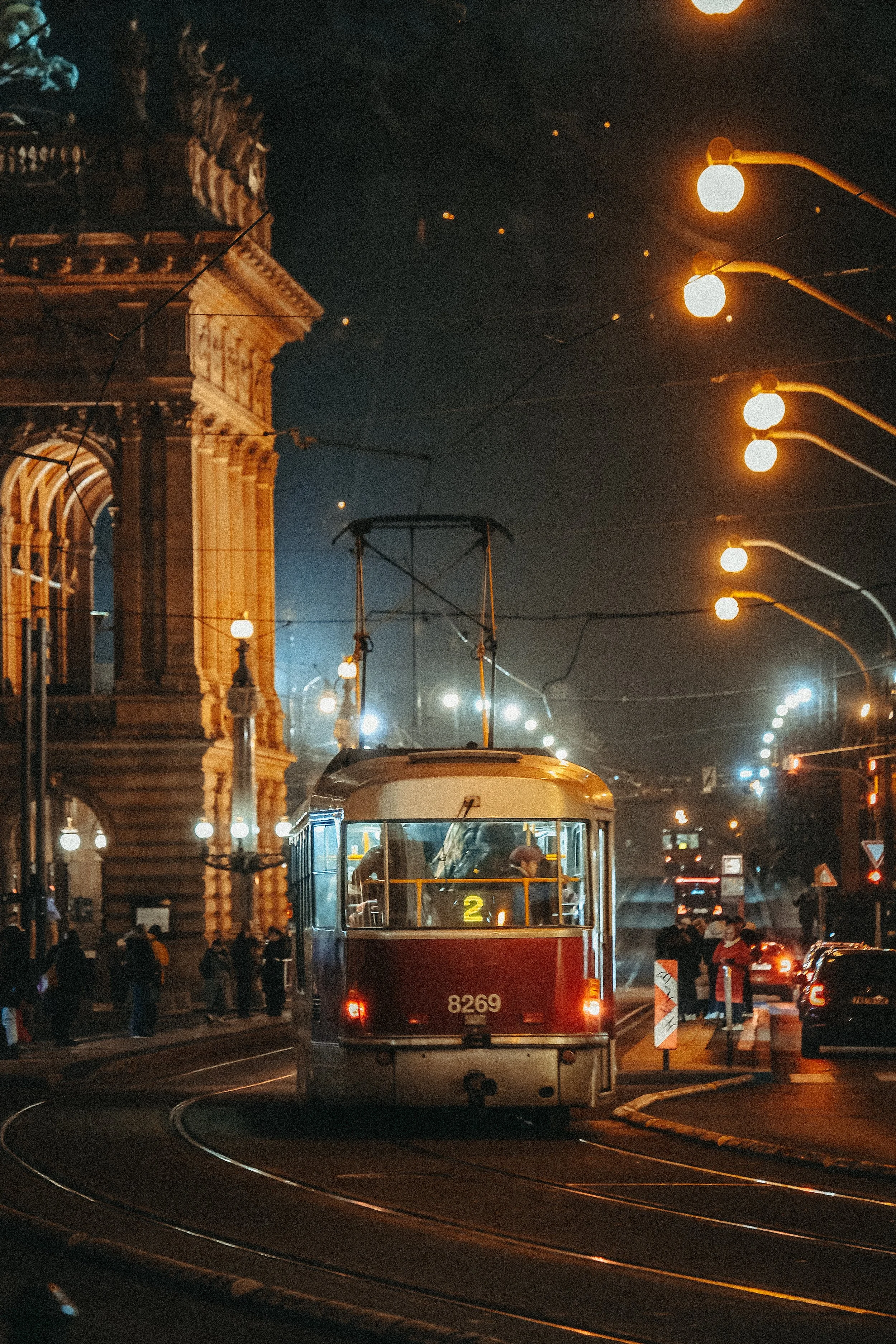 Straßenbahn fährt nachts an einem historischen Gebäude vorbei, umgeben von Straßenlaternen und Menschen auf dem Bürgersteig.