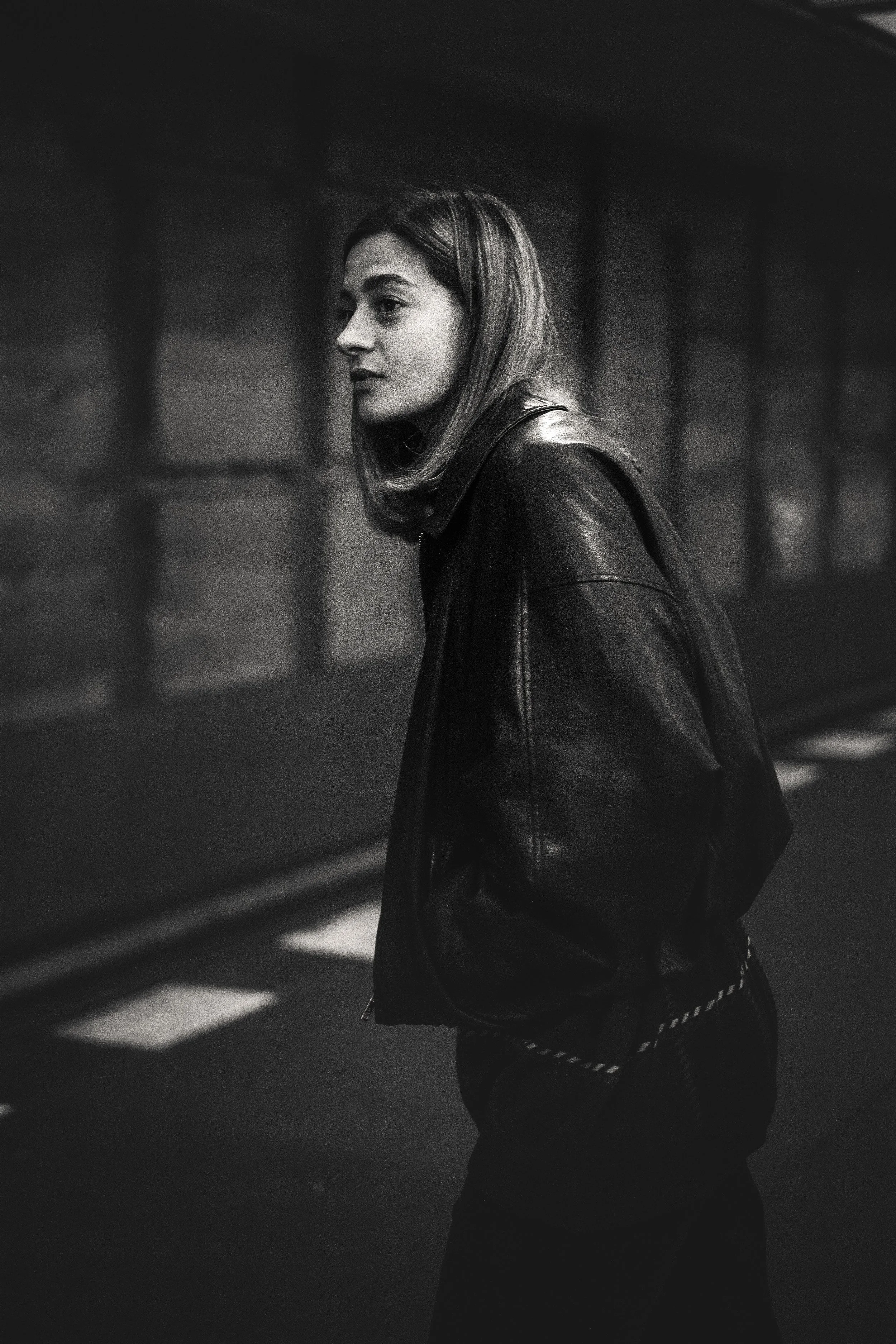 Schwarz-Weiß-Foto einer jungen Frau in Lederjacke, die auf der Straße steht, nach links blickt und nachdenklich wirkt.