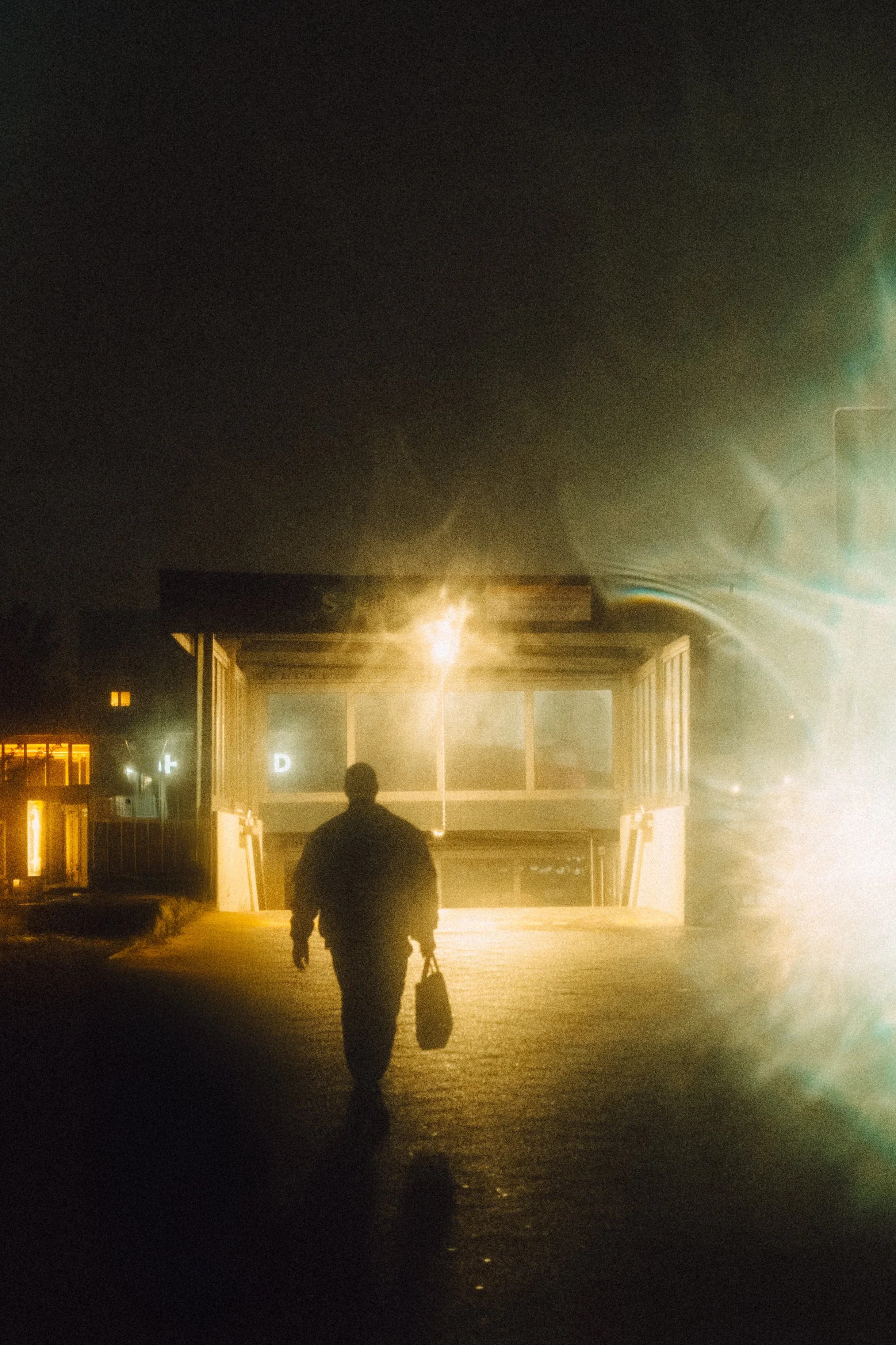 Ein Mann mit Aktentasche geht in der Nacht auf einen Bahnhofsvorplatz, beleuchtet von Straßenlaternen, mit einem futuristisch wirkenden Gebäude im Hintergrund.