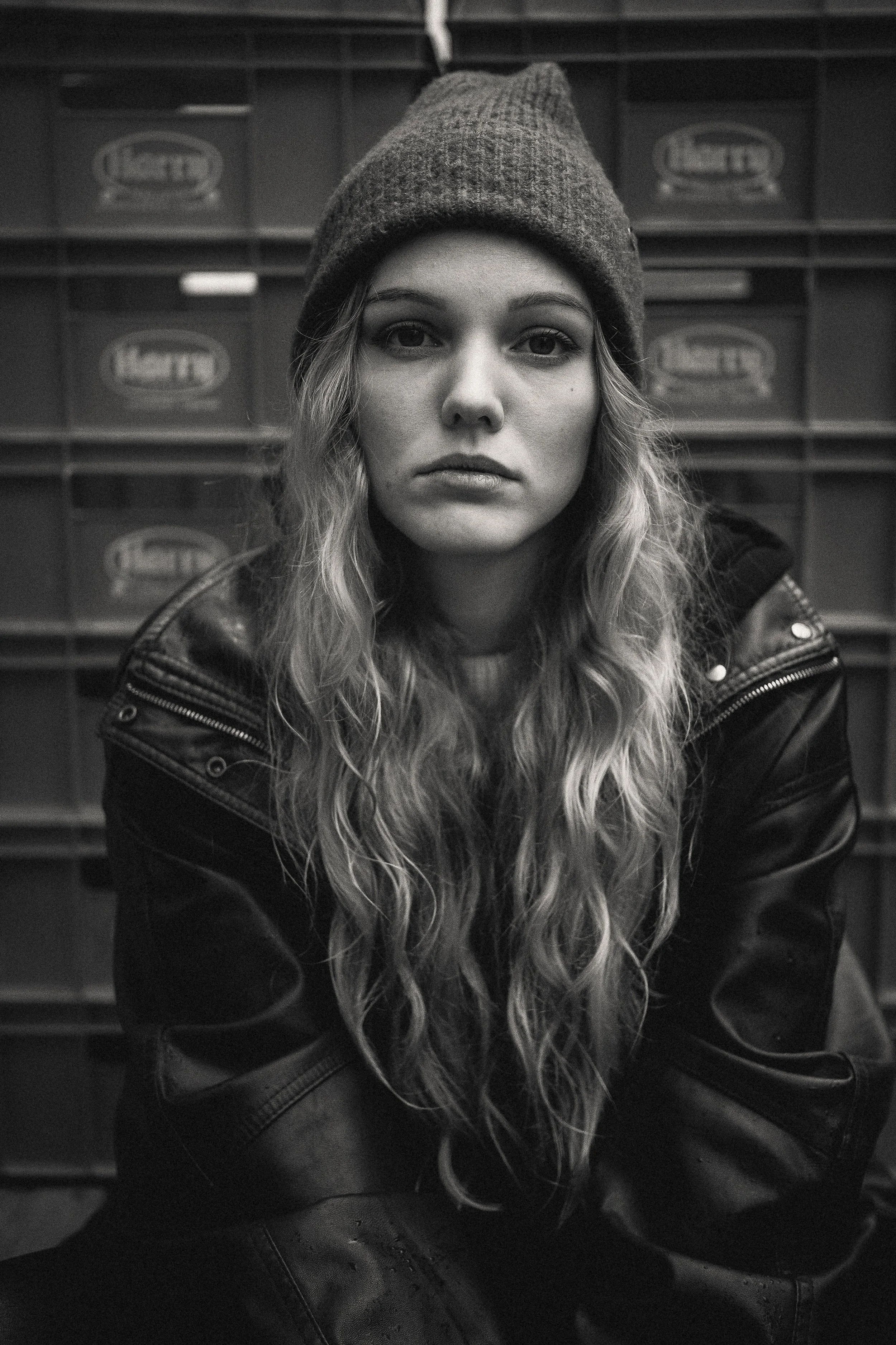 Schwarz-Weiß-Foto eines jungen Mädchens mit langen, gewellten Haaren, das eine Mütze trägt und eine Lederjacke trägt, vorne sitzend, mit einem Hintergrund aus Lagerkisten.