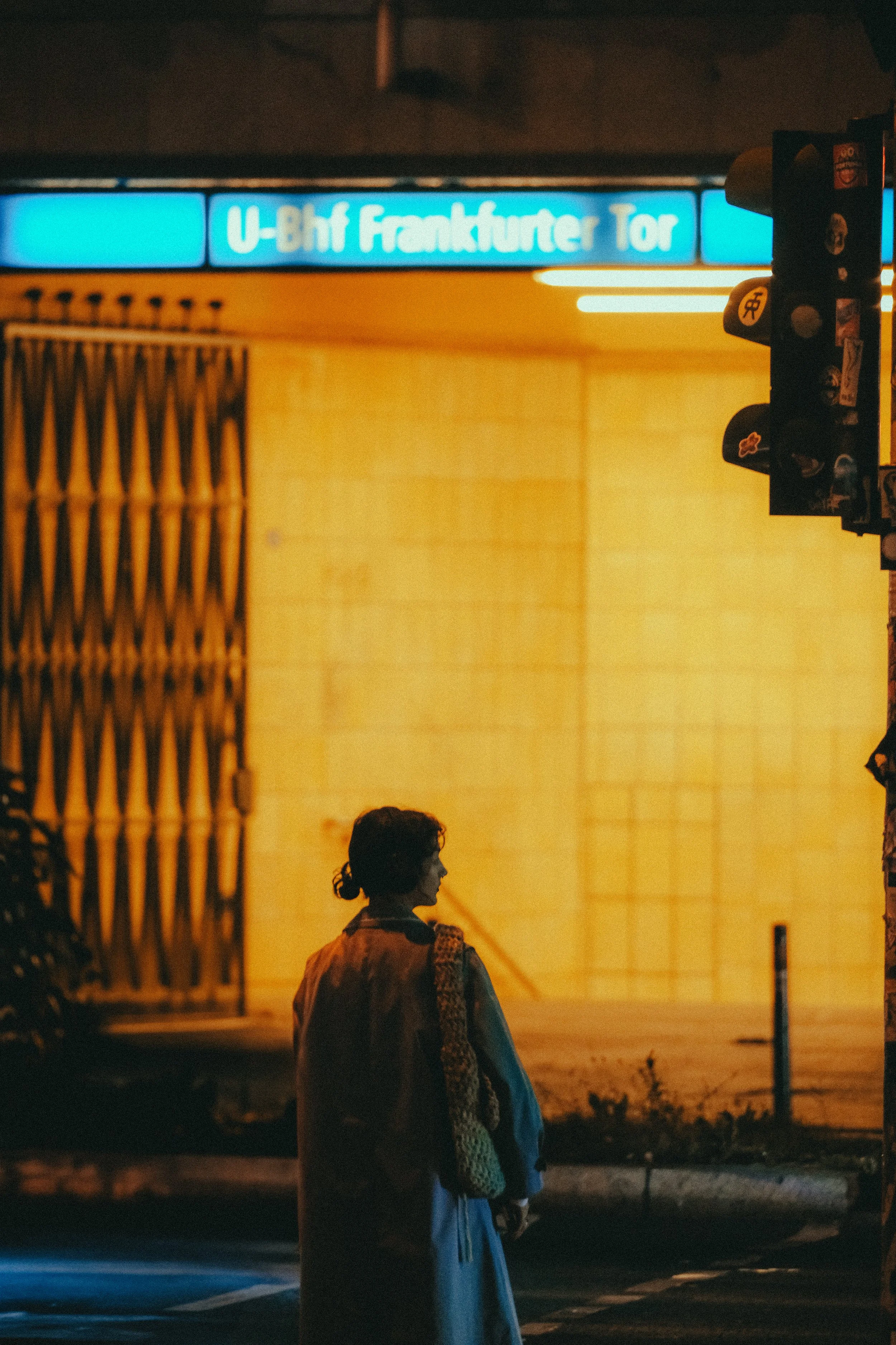 Eine Frau steht an einer Kreuzung bei Nacht, im Hintergrund leuchtendes orangefarbenes Licht und ein bläulich leuchtendes U-Bahn-Schild mit der Aufschrift 'U-Bhf Frankfurter Tor'.