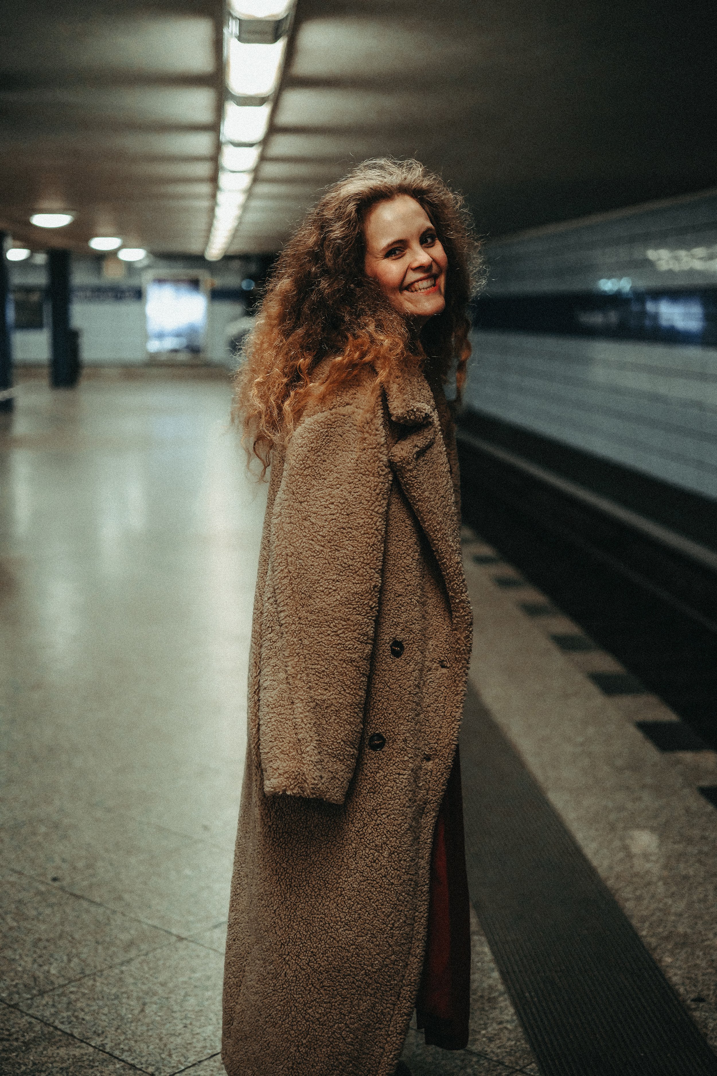 Junge Frau mit Locken in einem beige Mantel in einer U-Bahn-Station.