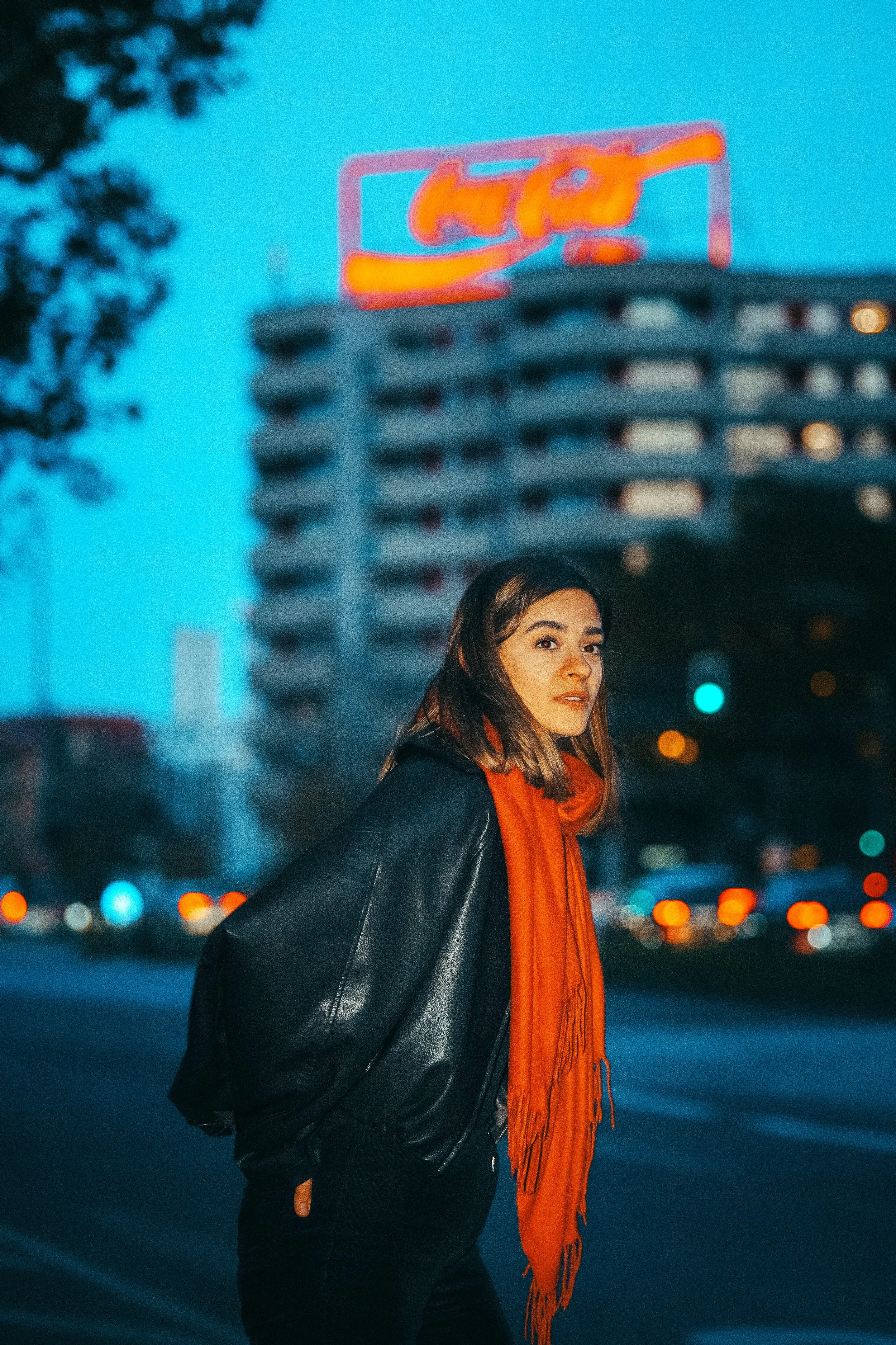 Eine junge Frau mit dunklem Haar, schwarzer Lederjacke und orangefarbenem Schal steht auf einer Straße bei Dämmerung, im Hintergrund sind bunte Lichter und ein mehrstöckiges Gebäude mit leuchtendem Neon-Schild.