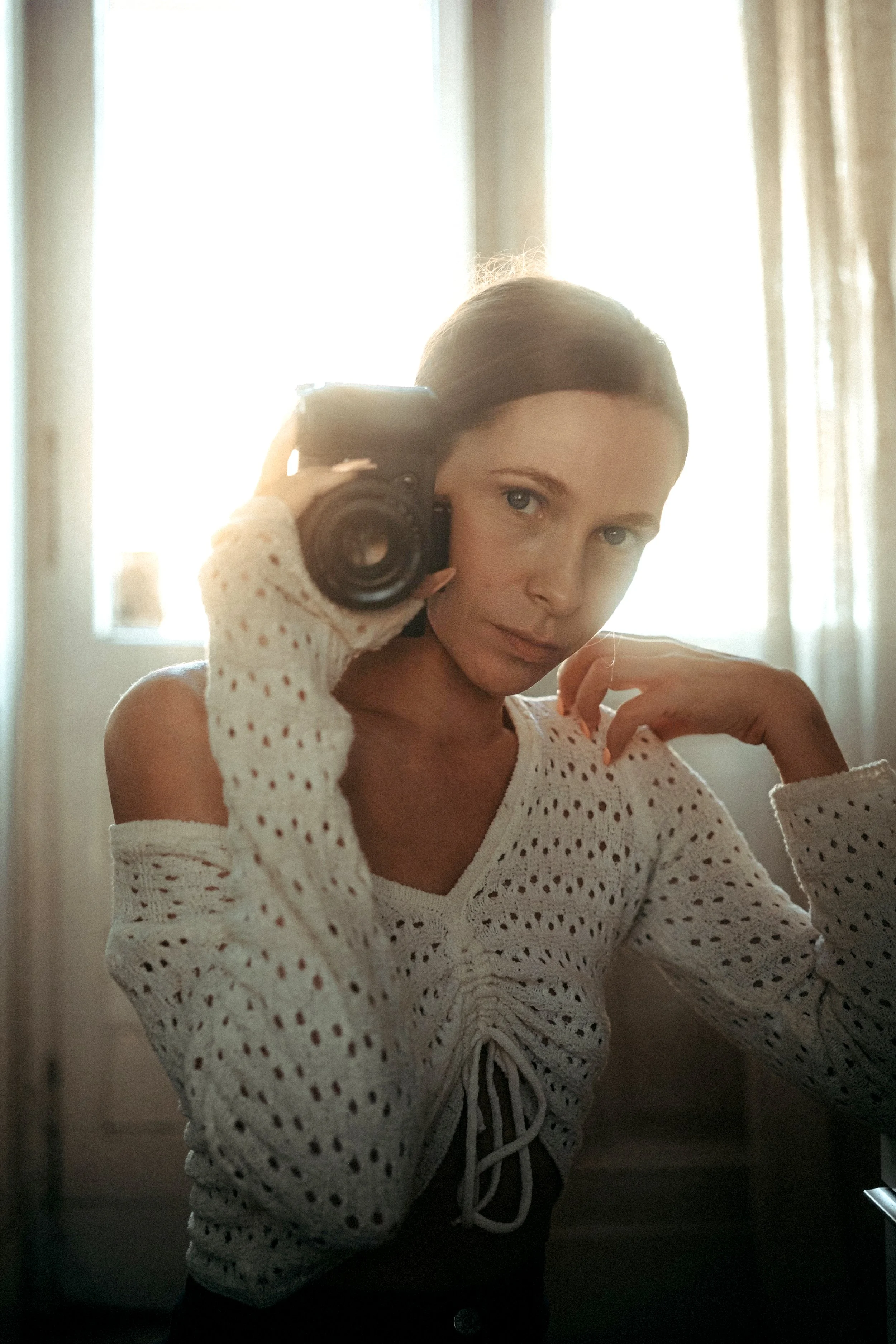 Junge Frau nimmt Selbstporträt im Spiegel mit Kamera auf, steht vor einem Fenster, trägt eine weiße, luftige Bluse mit Lochmuster, im warmen natürlichen Licht.