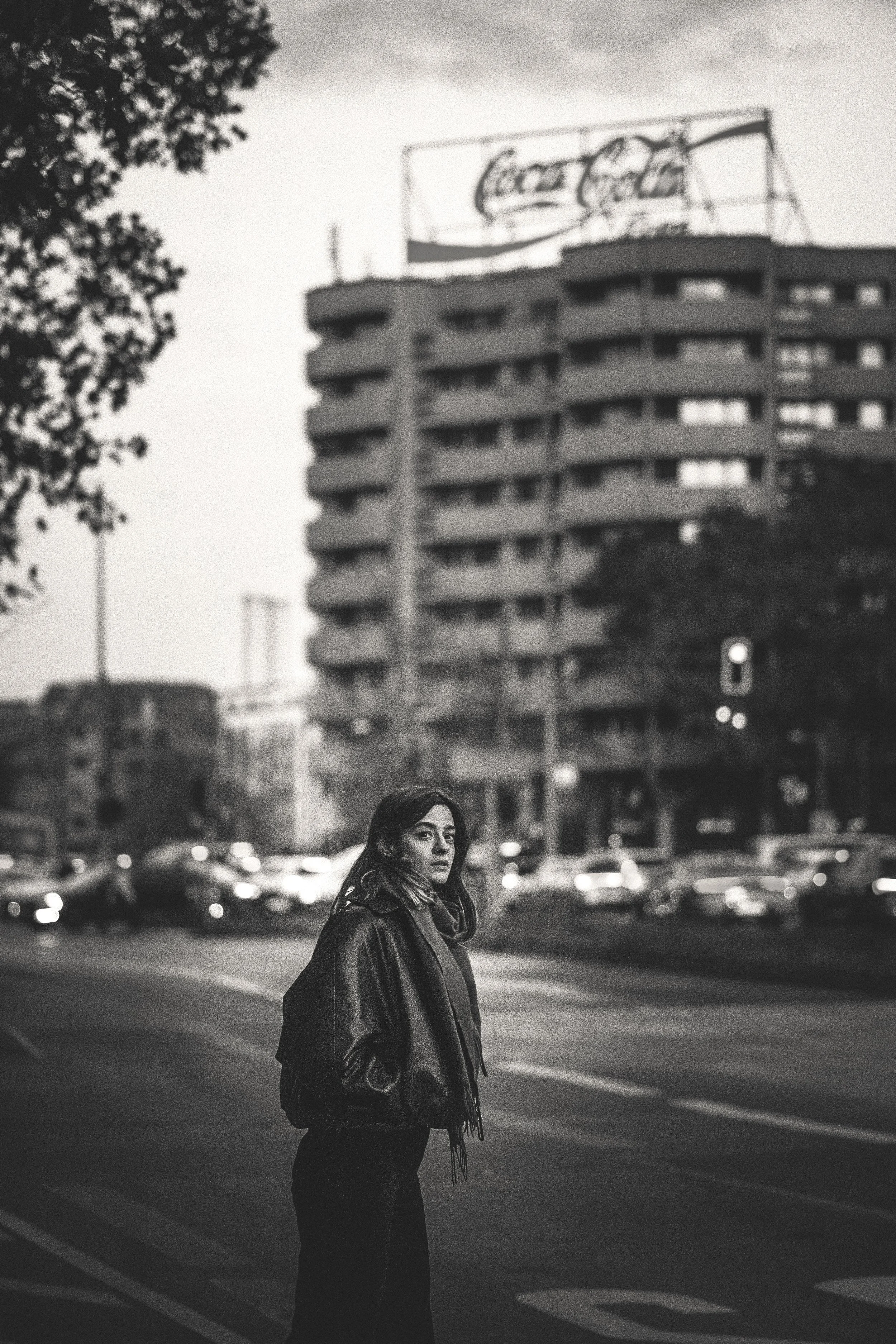 Eine junge Frau steht auf einer Straße in einer Stadt, umgeben von Autos und einem hohen Gebäude im Hintergrund, in Schwarz-Weiß aufgenommen.