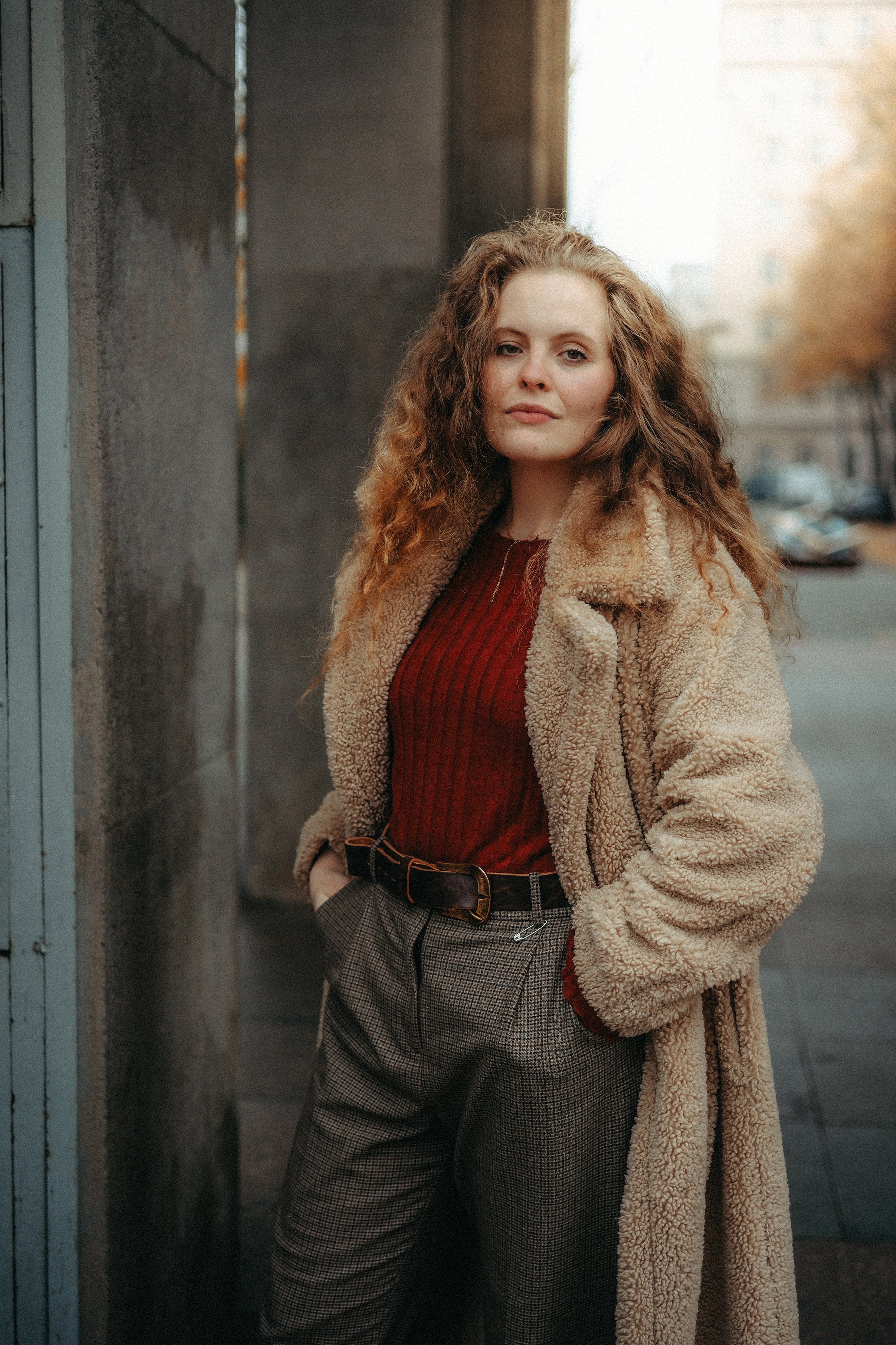 Eine junge Frau mit lockigem, braunem Haar steht auf einer Stadtstraße, trägt einen braunen Teddyfellmantel, eine rote Strickjacke und karierte Hosen.