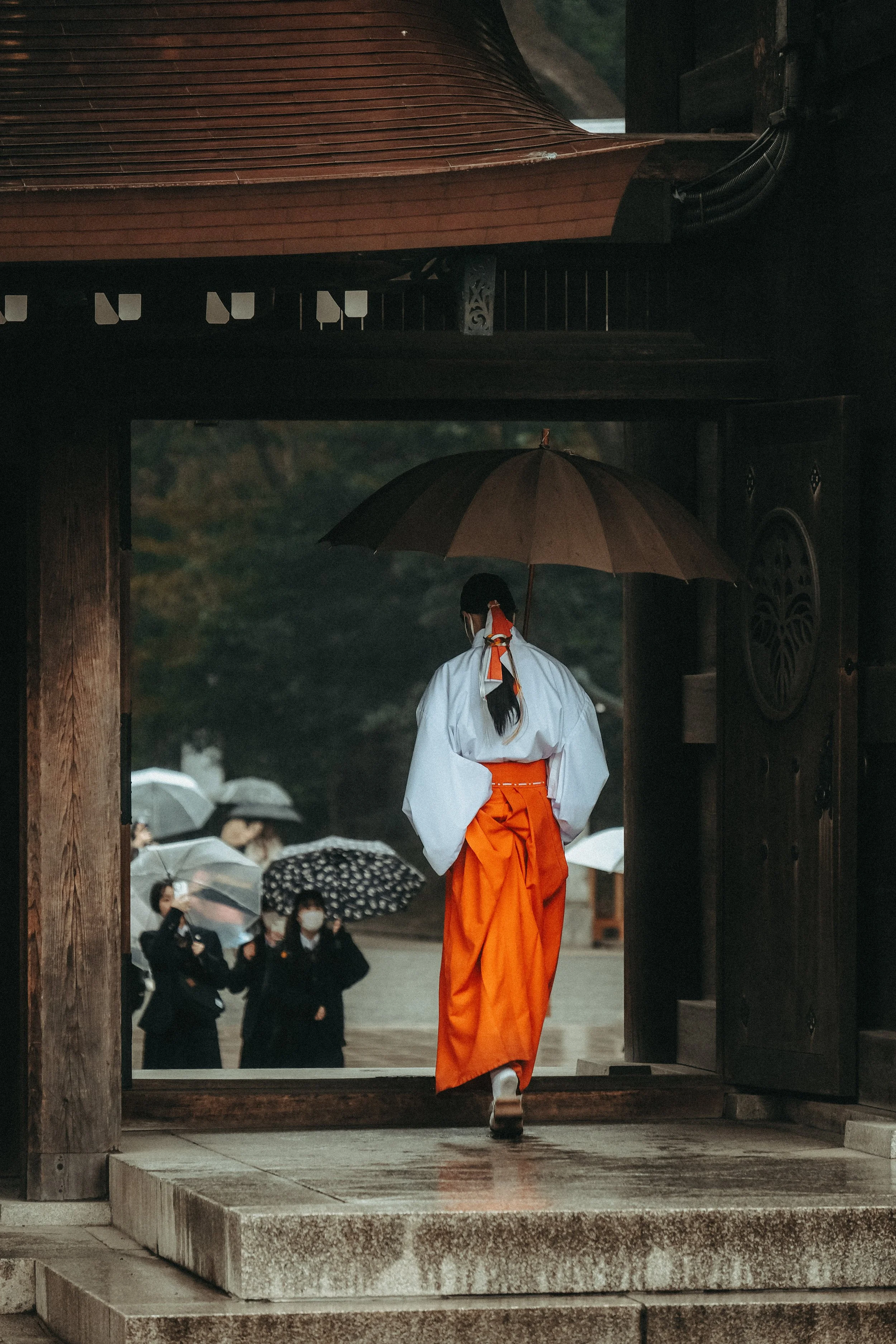 Eine Person in traditioneller japanischer Tracht, die durch ein Tor schreitet, im Regen, während mehrere Menschen mit Regenschirmen im Hintergrund stehen.