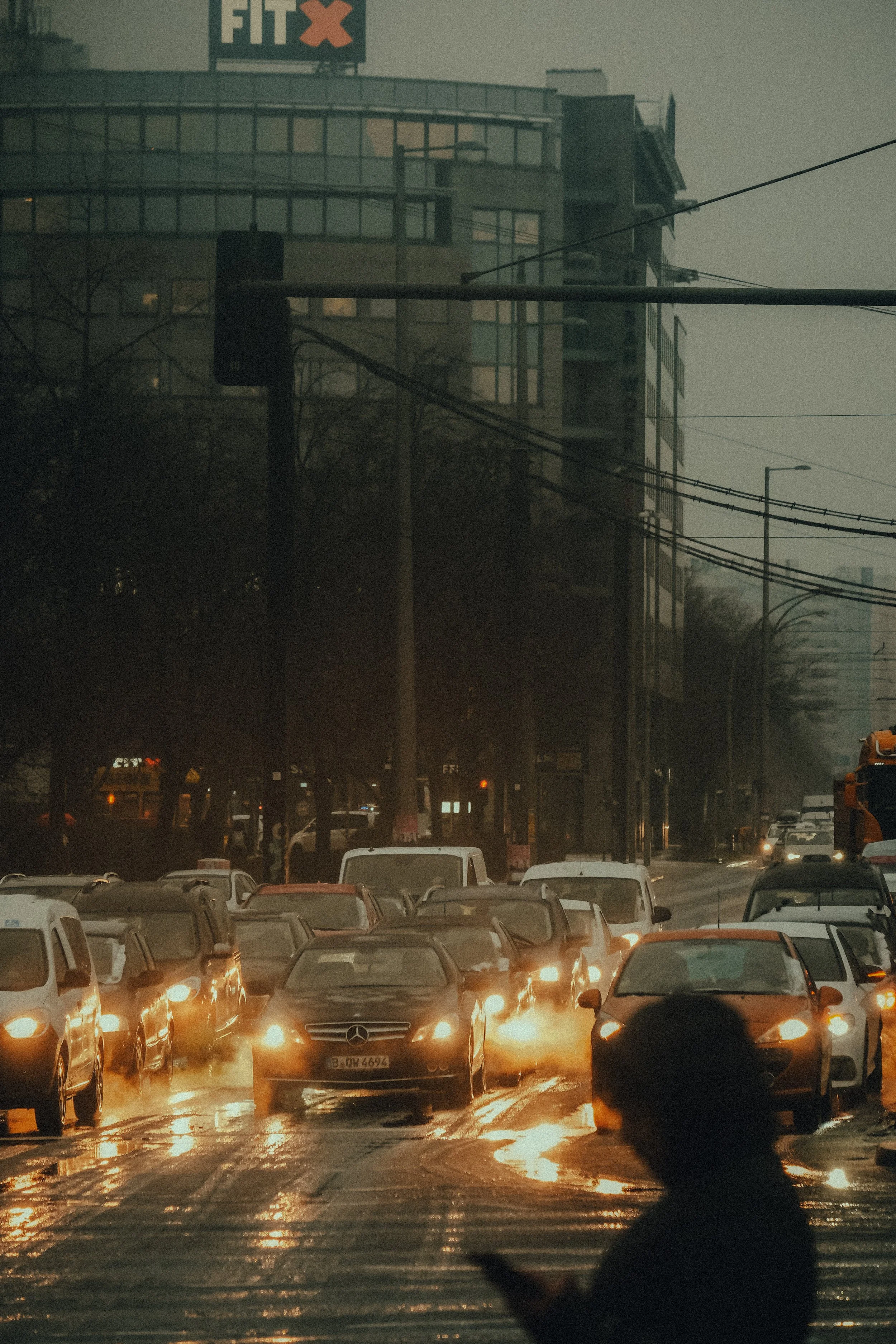 Stau auf einer Stadtstraße bei Regen mit reflektierenden Wasserpfützen, dunkler Himmel und im Vordergrund eine Person, die auf ihr Handy schaut.