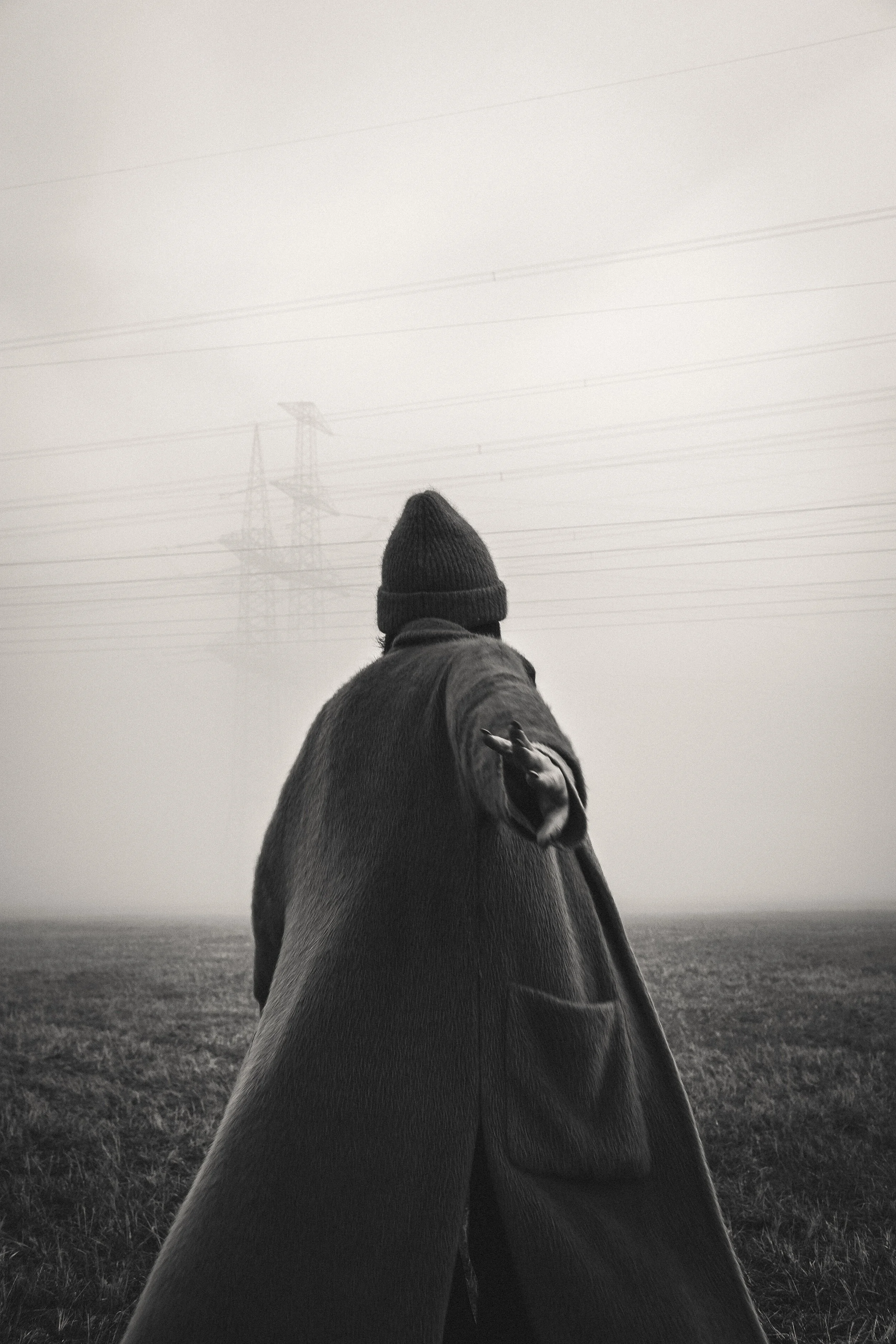 Schwarz-Weiß-Foto eines Menschen in einem langen Mantel und einer Kappe, der auf einem Feld steht und in die Ferne blickt, mit Hochspannungsleitungen im Hintergrund.