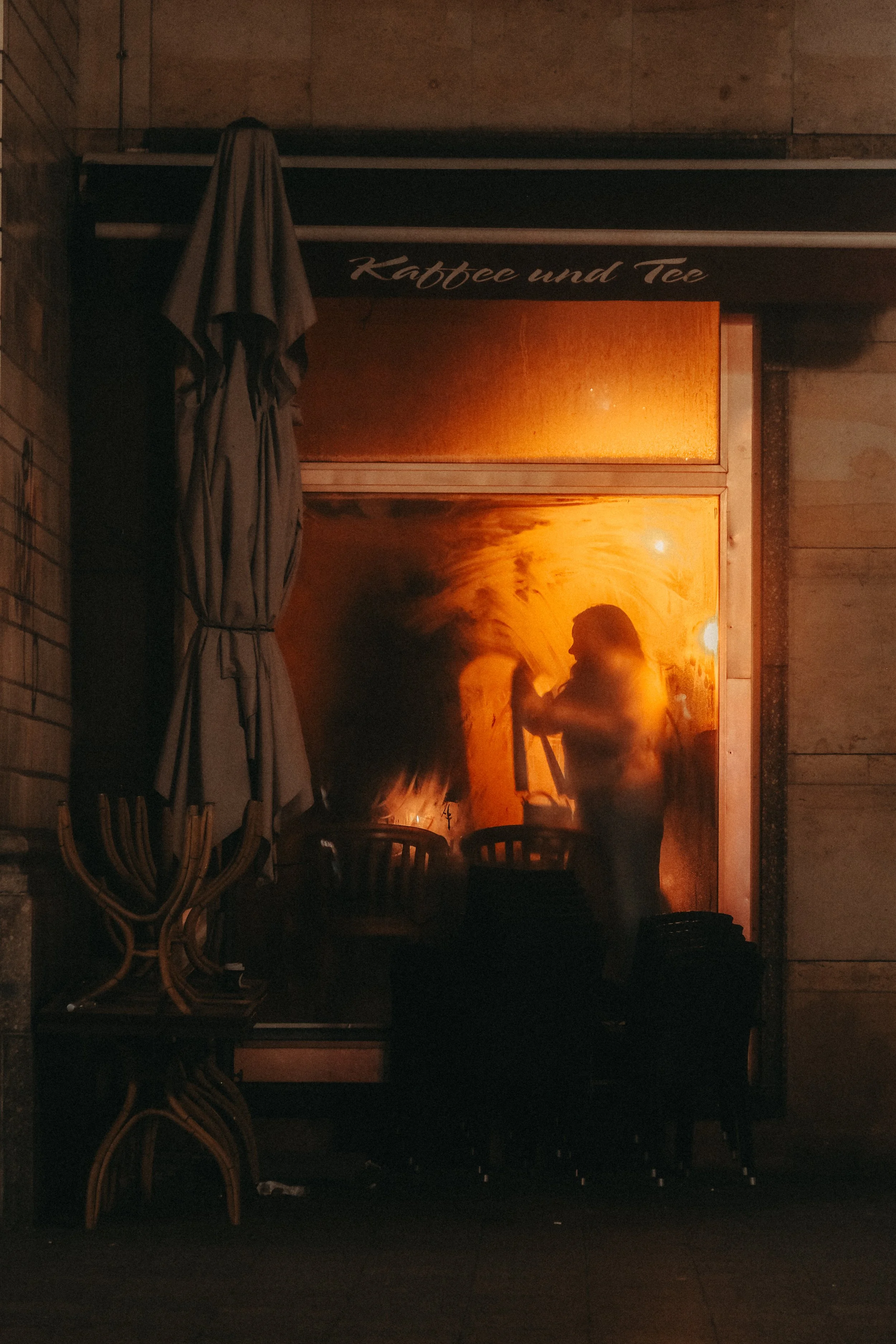 Schatten einer Person, die durch ein warm erleuchtetes Fenster eines Cafés namens 'Kaffee und Tee' schaut, mit Stühlen im Vordergrund.