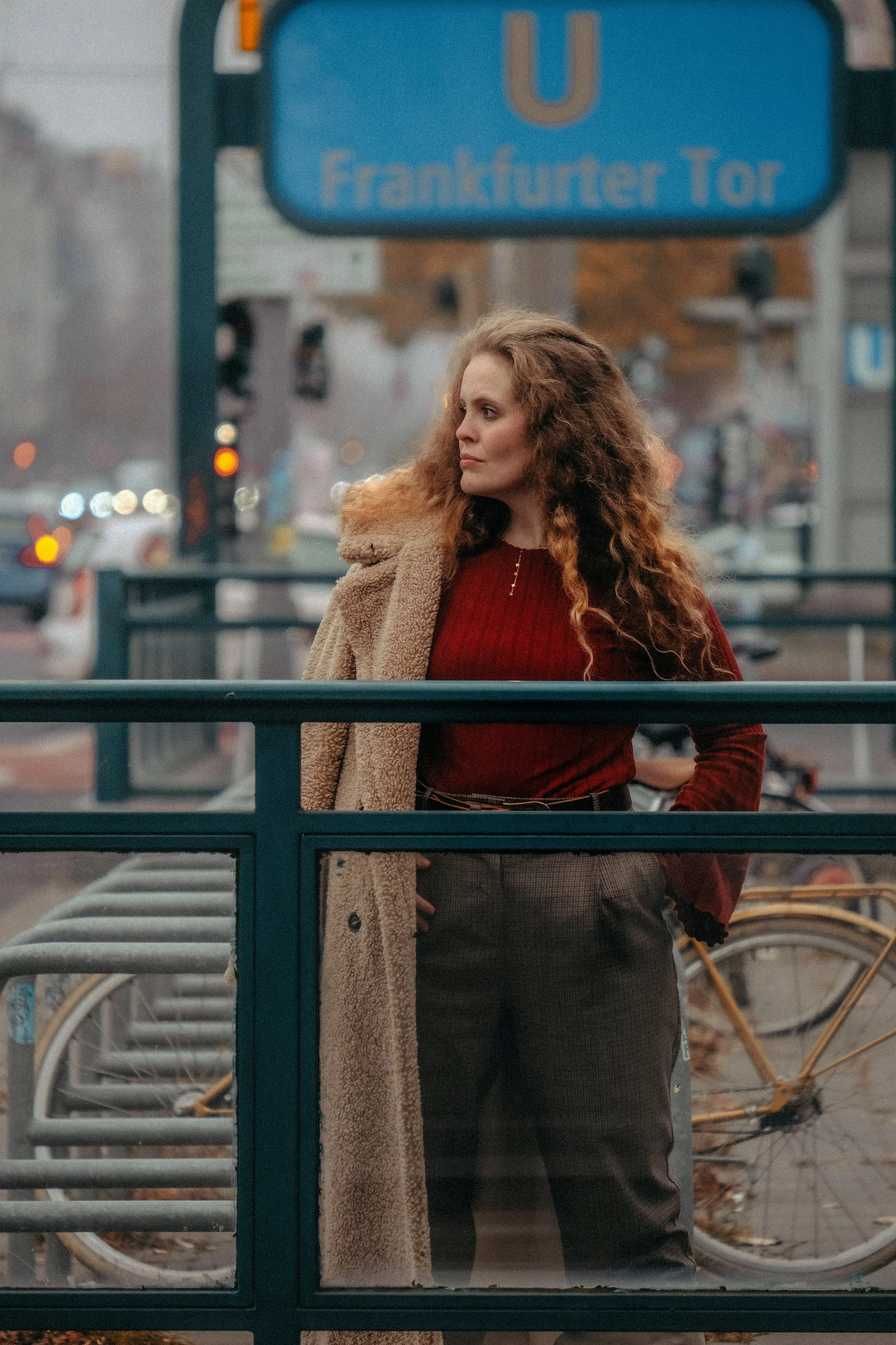 Eine Frau mit lockigem Haar steht an einem U-Bahnhof in Frankfurt, schaut nach links, trägt einen roten Pullover und eine beige Jacke, im Hintergrund Fahrräder und Verkehr auf der Straße.