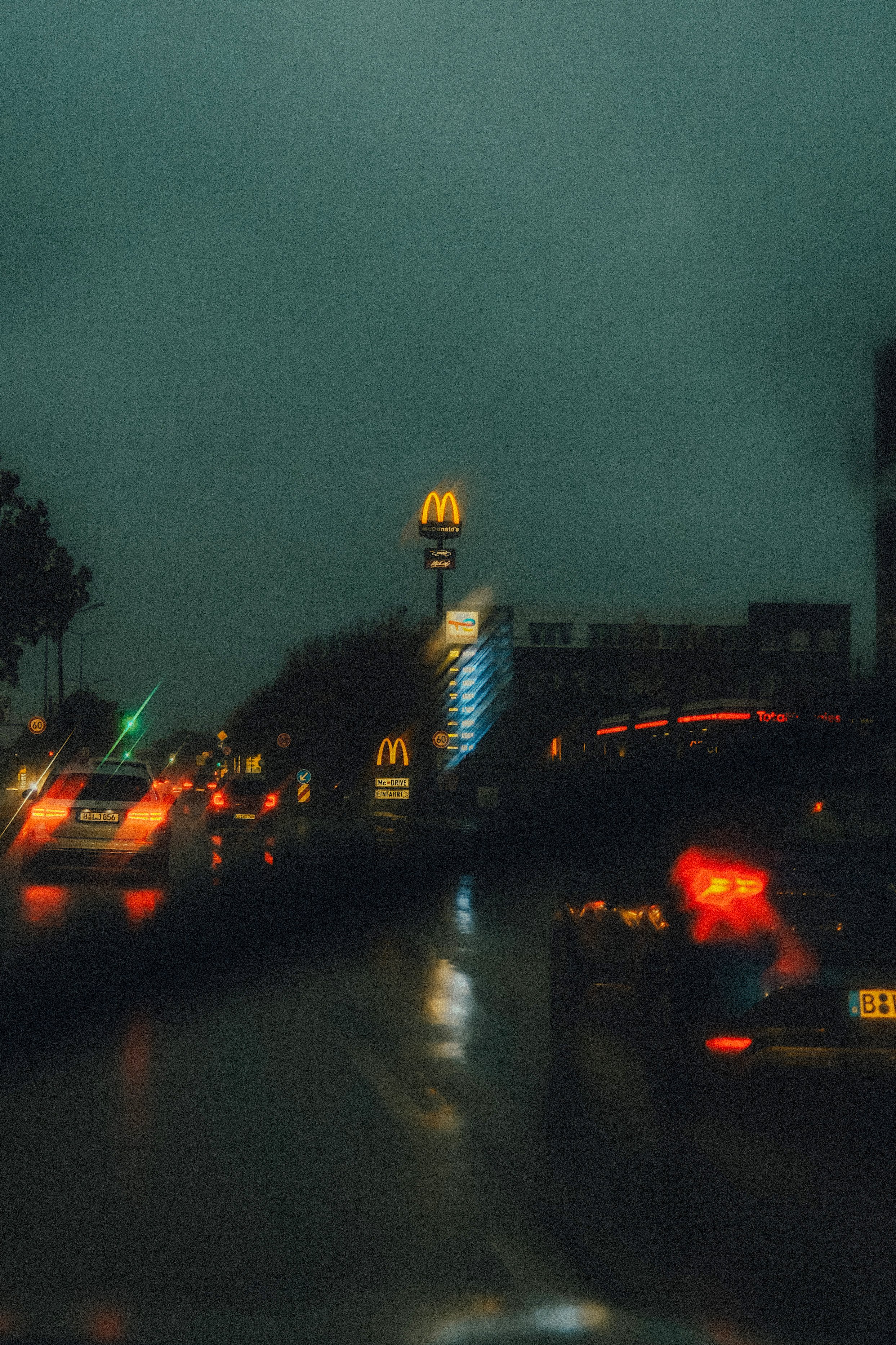 Nachtaufnahme einer stark befahrenen Straße mit nassen Straßen, reflektierenden Lichtern und einem McDonald's Schild im Hintergrund.