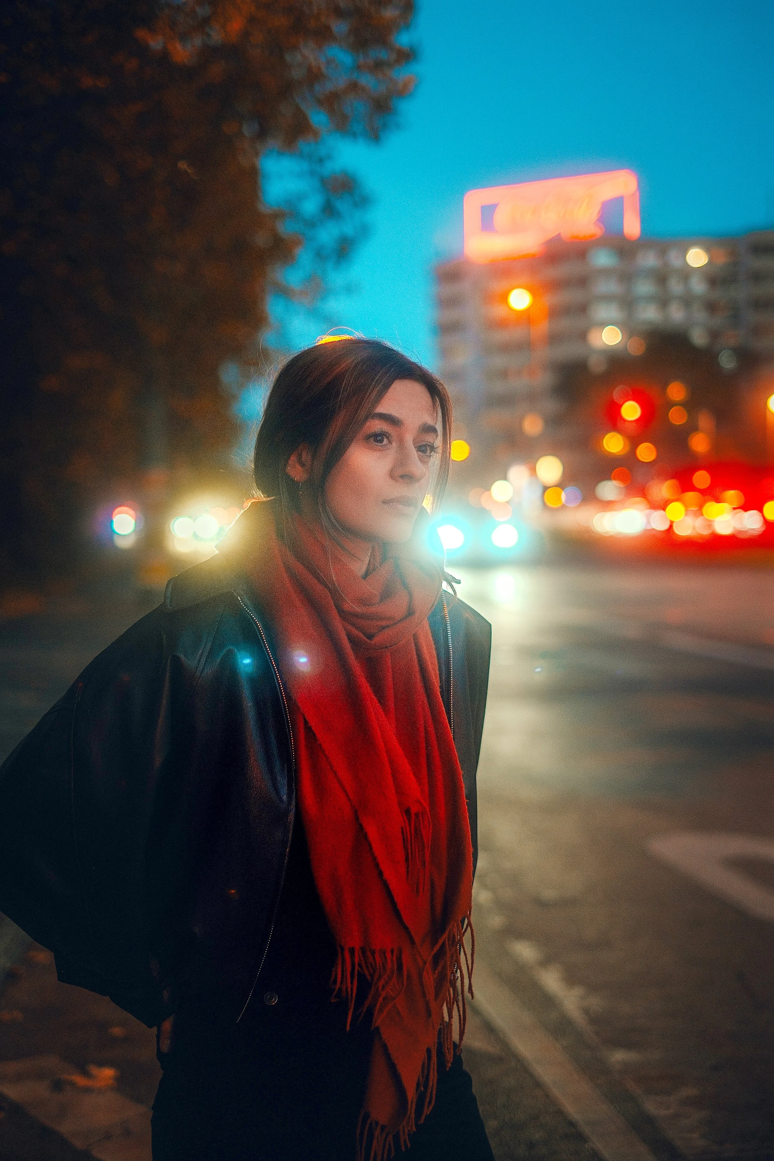 Junge Frau steht nachts an einer belebten Straßenkreuzung, trägt eine schwarze Lederjacke und einen roten Schal, im Hintergrund leuchtende Neonlichter und Gebäude.