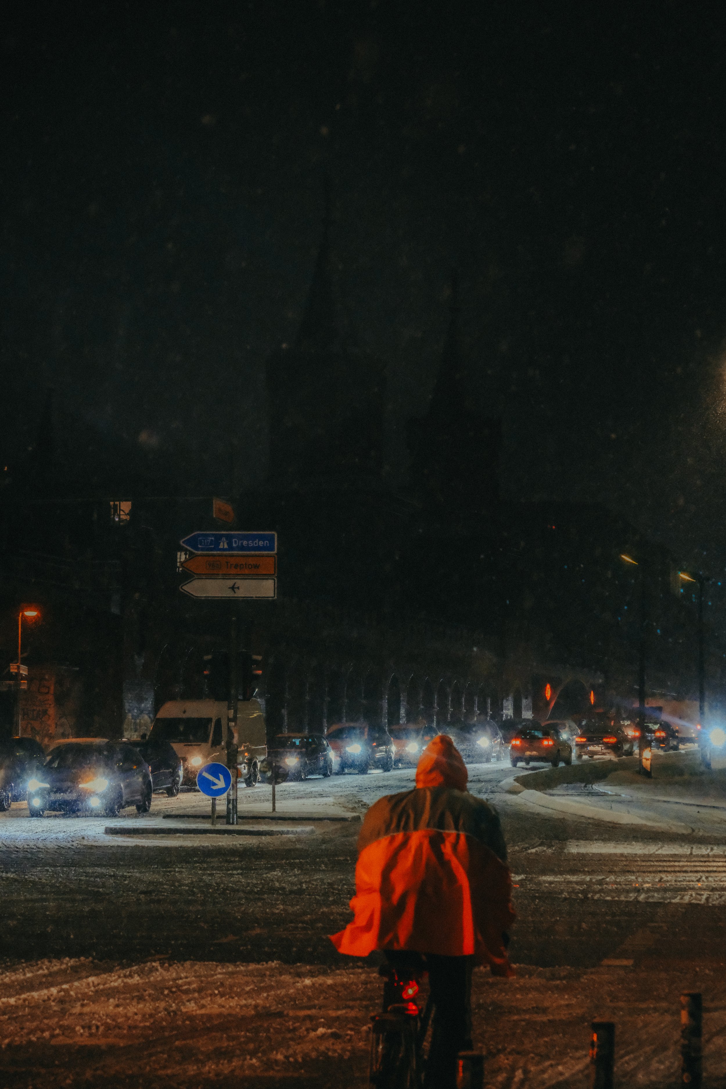 Eine einzelne Person fährt nachts auf einem Fahrrad, eingekuschelt in eine orangefarbene Jacke, während im Hintergrund Autos auf einer Straße in Dresden unterwegs sind, bei Schneefall und Dunkelheit.