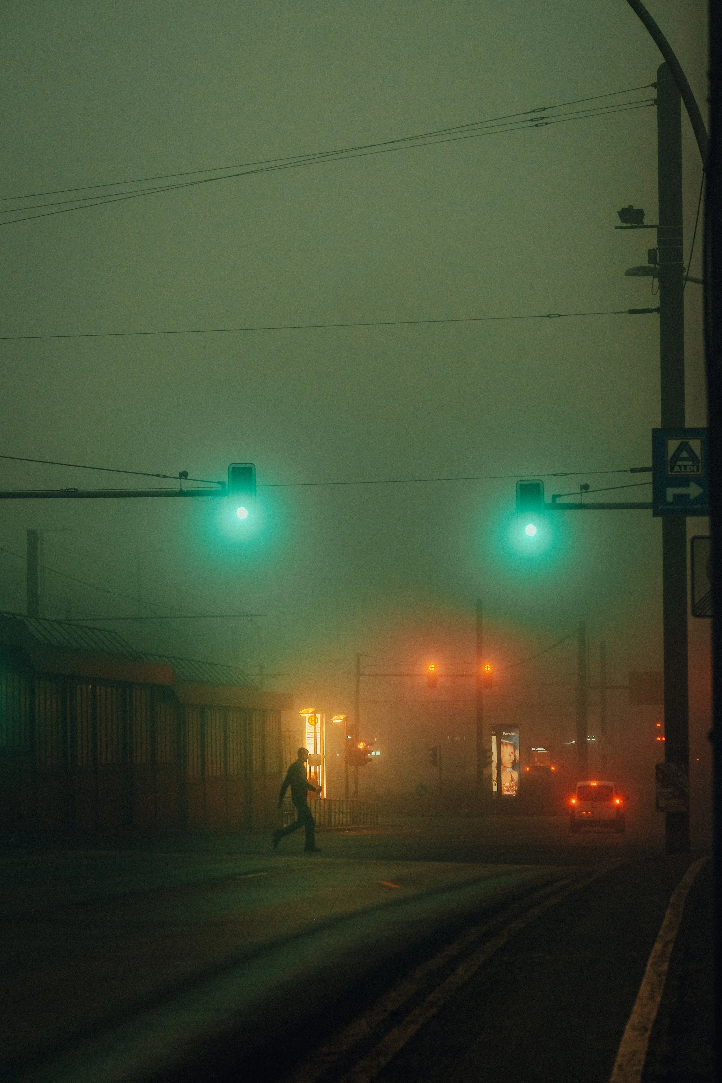 Nebelverhangene Straße bei Nacht mit grünen Ampeln und einem Fußgänger.