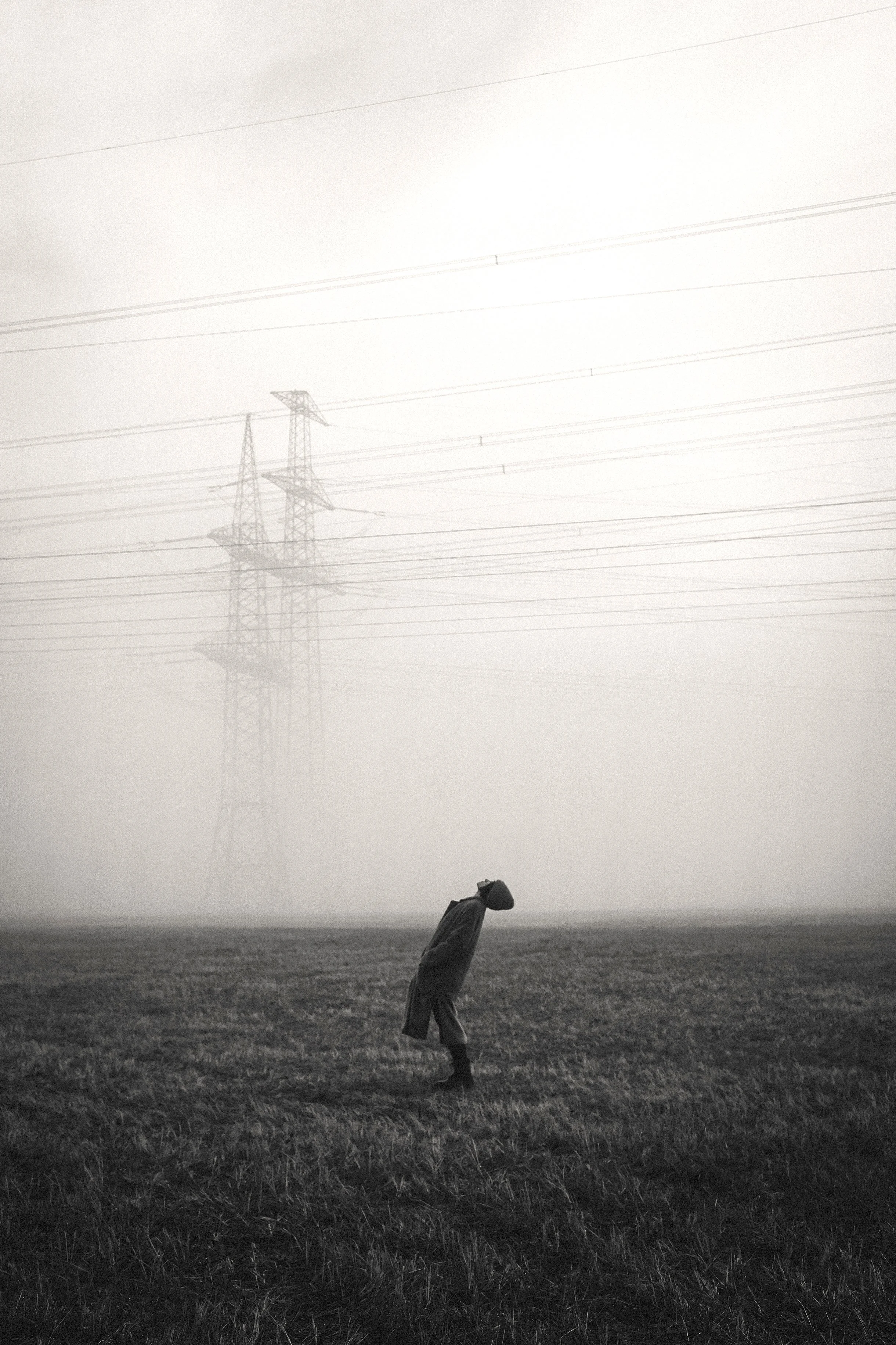 Eine einzelne Person steht auf einem grasbewachsenen Feld im Nebel, mit Hochspannungsleitungen im Hintergrund.