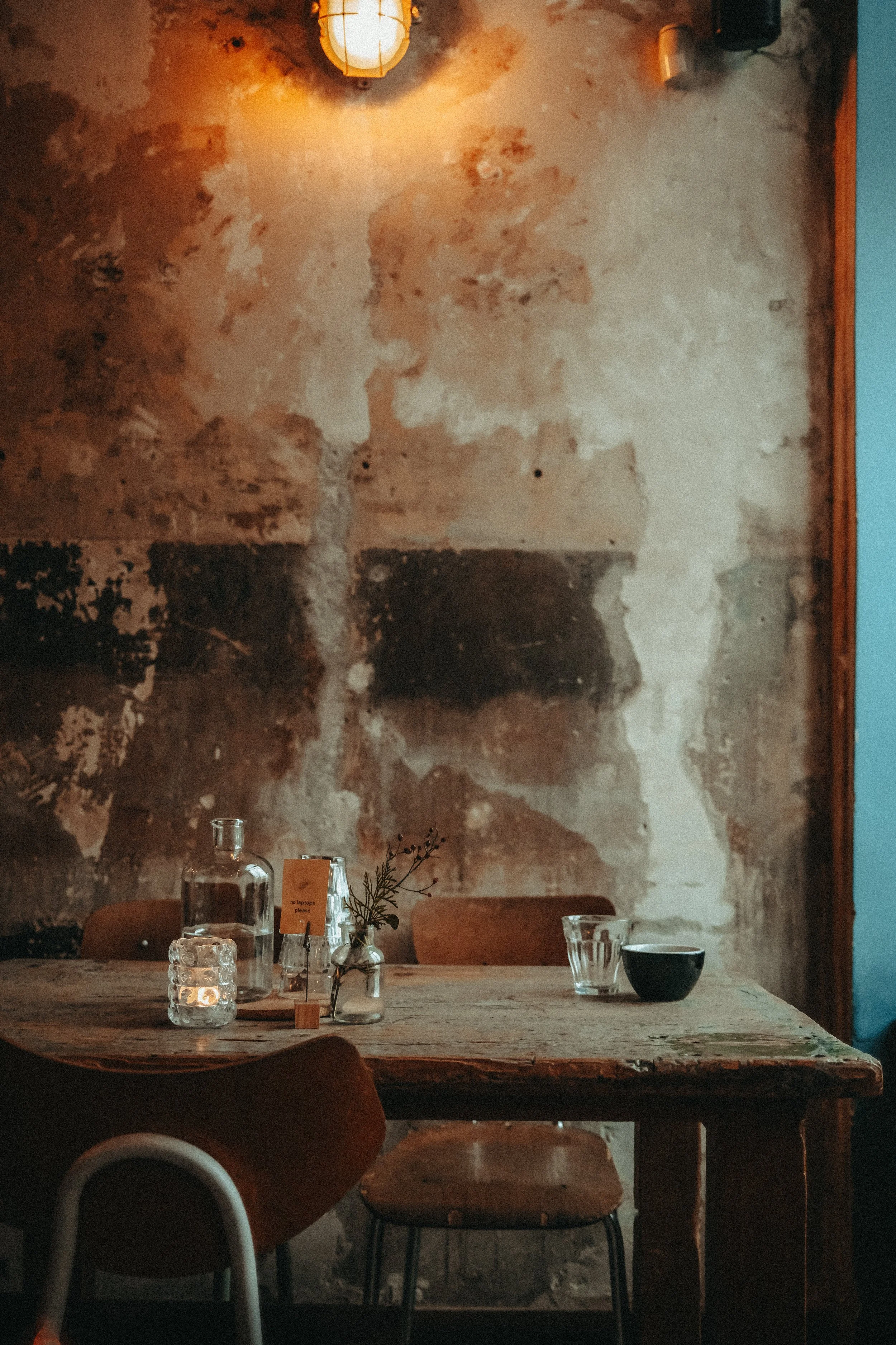 Rustikaler Esstisch in einem stilvollen Café mit Kerzen, einer Glaskaraffe, Gläsern, einer Vase mit Zweigen und einer schwarzen Schüssel, vor einer alten, abgenutzten Wand.