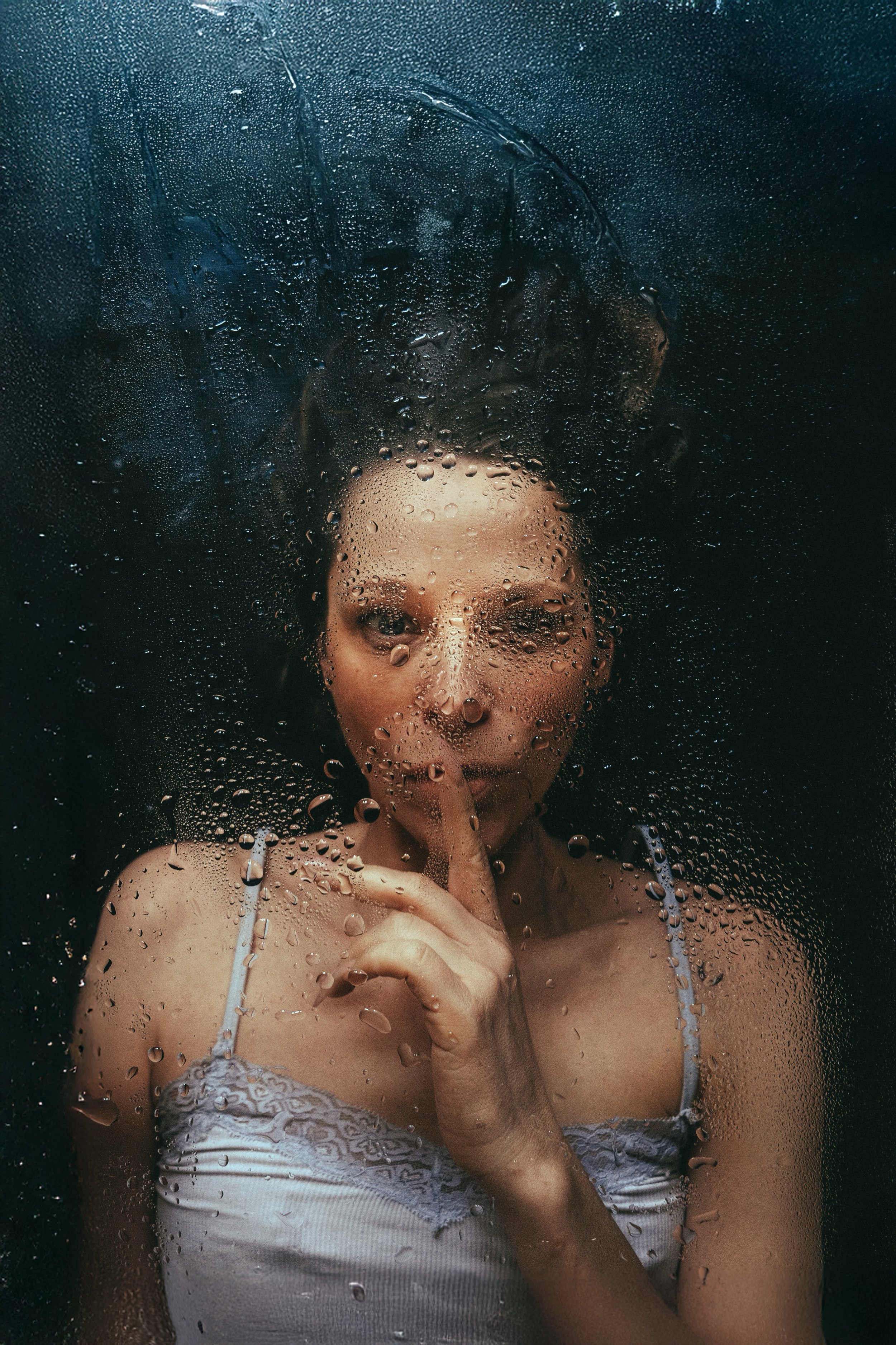 Frau hinter einer Glasscheibe mit Wassertröpfchen, die einen Finger vor den Mund hält.