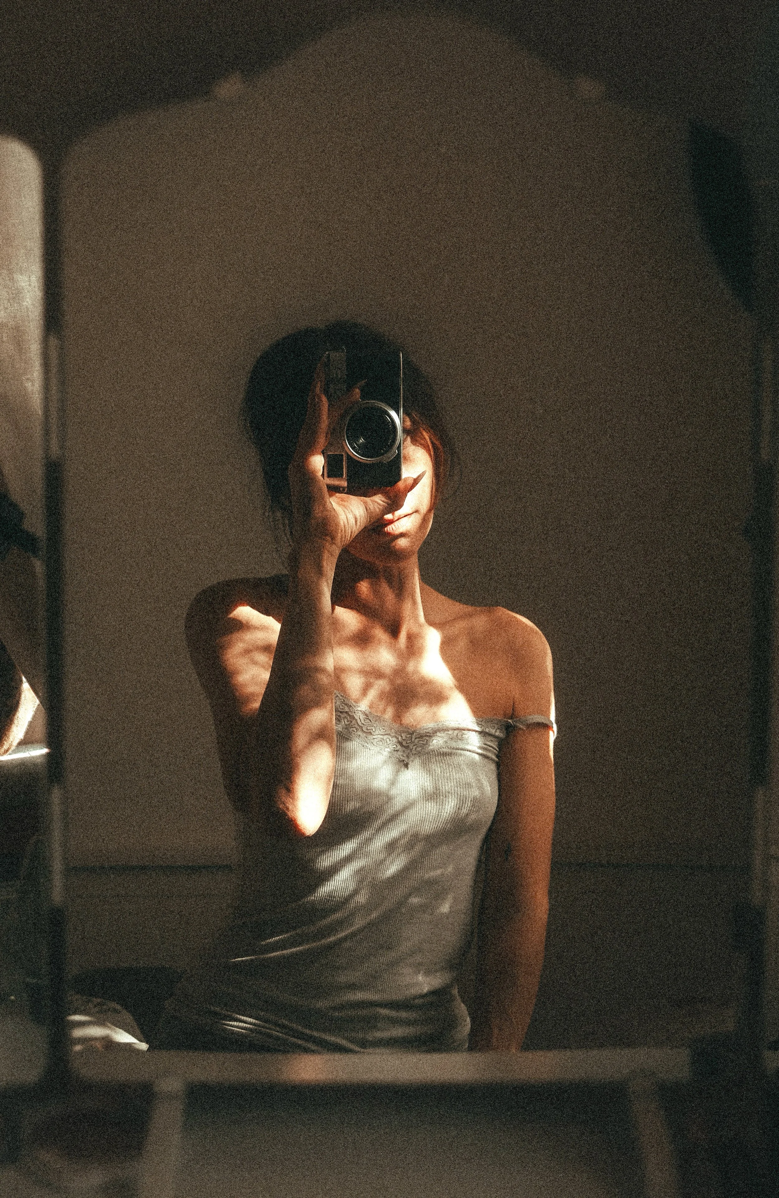 Eine Frau nimmt ein Selfie vor einem Spiegel auf, bei dem sie nur einen Teil ihres Gesichts und ihre Schultern sichtbar ist. Die Beleuchtung ist warm und das Bild hat einen künstlerischen, intimen Stil.