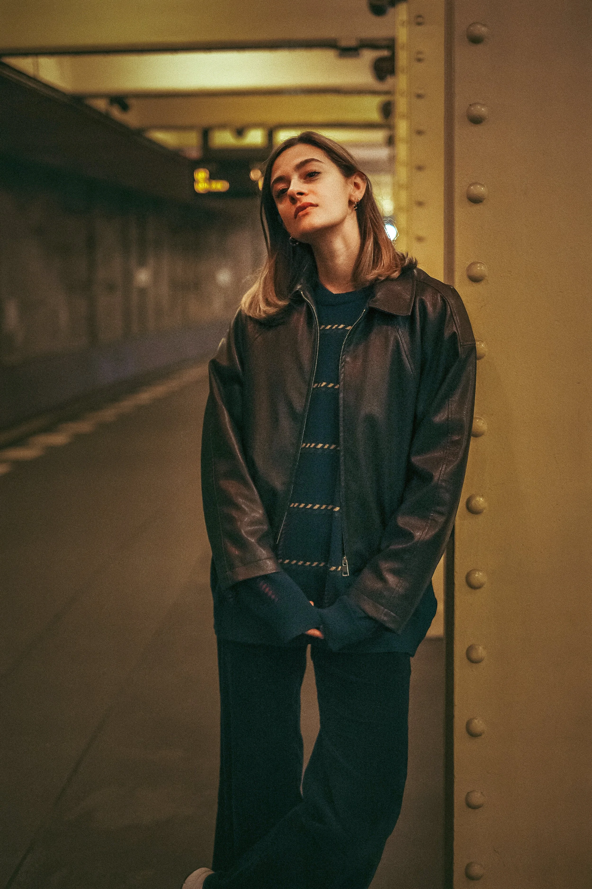 Junge Frau steht in einem Tunnel, trägt eine braune Lederjacke und schwarze Pants, und lehnt an einer gelben Stützsäule.