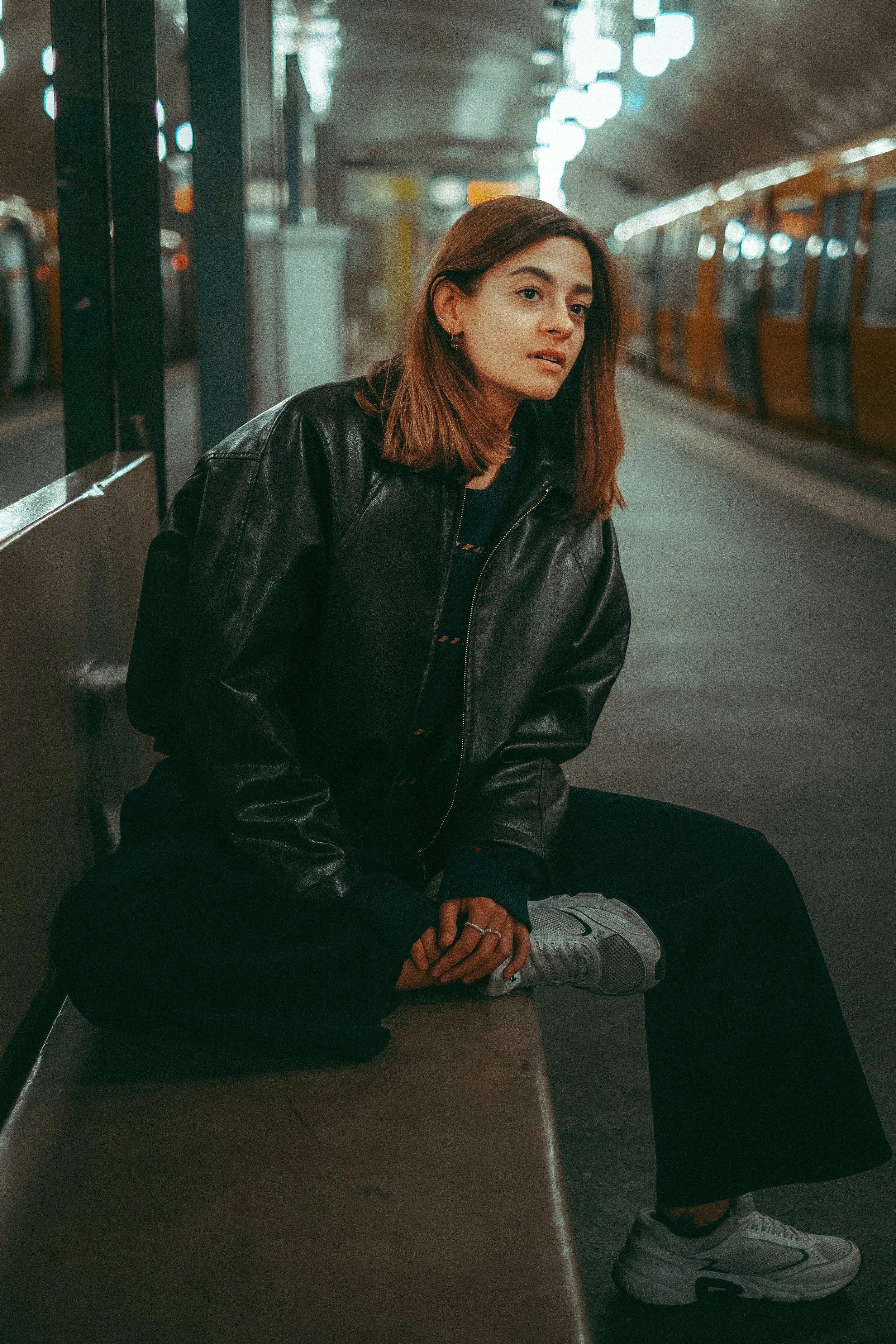 Eine junge Frau sitzt auf einer Bank in einem U-Bahnhof, trägt eine schwarze Lederjacke, dunkle Hosen, weiße Sneakers und hat braune, schulterlange Haare.
